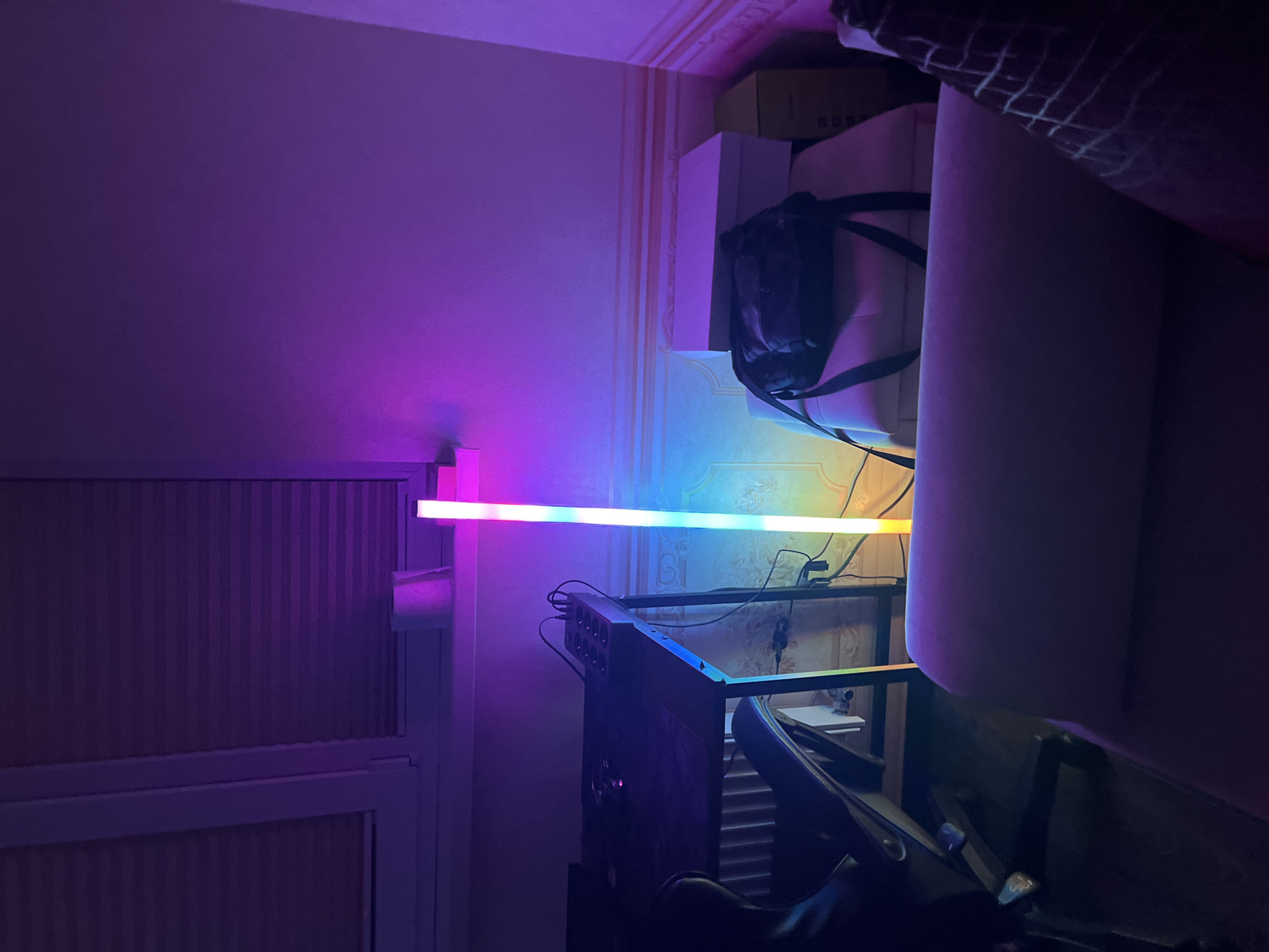 In an indoor scene illuminated by colorful LED lighting, a long, slender LED light bar stands vertically in the center of the frame, displaying a gradient of colors from yellow at the bottom to pink at the top. To the left, a portion of a dark-colored desk is visible, with an office chair in front of it. On the desk, various electronic devices and cables are present. Behind the desk, a white radiator can be seen. To the right of the LED light bar, a black backpack rests on top of a white box, which is stacked on another flat surface. Further to the right, a cardboard box is positioned vertically. In the foreground, the edge of a large piece of furniture, possibly a sofa, is visible, covered in a light-colored fabric that reflects the ambient lighting. The walls are decorated with patterned wallpaper. The environment appears to be a room within a residential building. The specific location in Lviv, Ukraine, cannot be determined from the image.