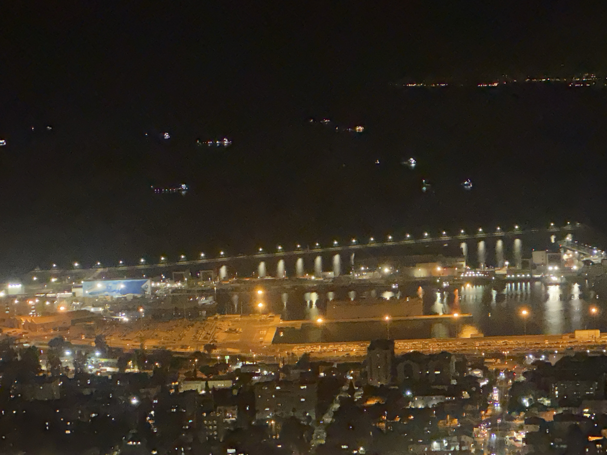 The image depicts a nighttime aerial view of the port of Haifa, Israel. The scene is dominated by the dark water of the bay, illuminated by the lights of ships, docks, and the city. Several large cargo ships and smaller vessels are visible in the harbor, their lights reflecting on the water. The port facilities, including cranes and loading areas, are brightly lit. In the foreground, the city of Haifa is seen with numerous illuminated buildings and streetlights. The lights stretch along the coastline and inland, indicating a densely populated urban area. The overall impression is one of a bustling port city at night.