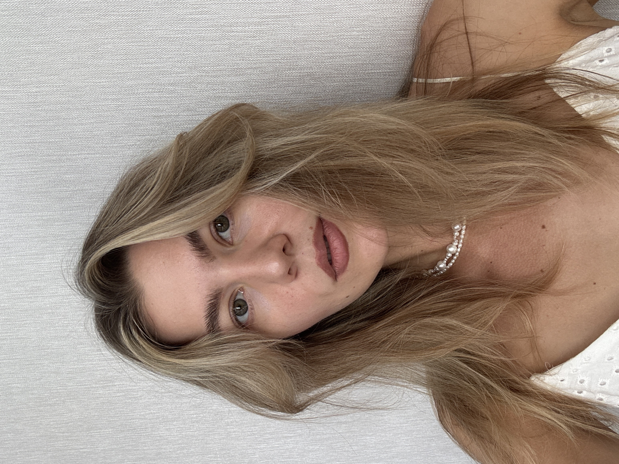 A close-up, head-and-shoulders shot captures a young woman with blonde hair, parted in the middle and styled with loose waves. Her head is tilted slightly to her left, and she is looking directly at the camera. She has fair skin, prominent cheekbones, and light green eyes. Her eyebrows are dark and full, and she is wearing light makeup, including lip gloss. A delicate pearl necklace adorns her neck, and she is wearing a white garment with eyelet detailing. The background is a textured light gray wall. The lighting is soft and even, suggesting it may be natural light from a window. The image appears to be taken indoors, likely in a residential setting. No specific location in a city is identifiable from the provided image.