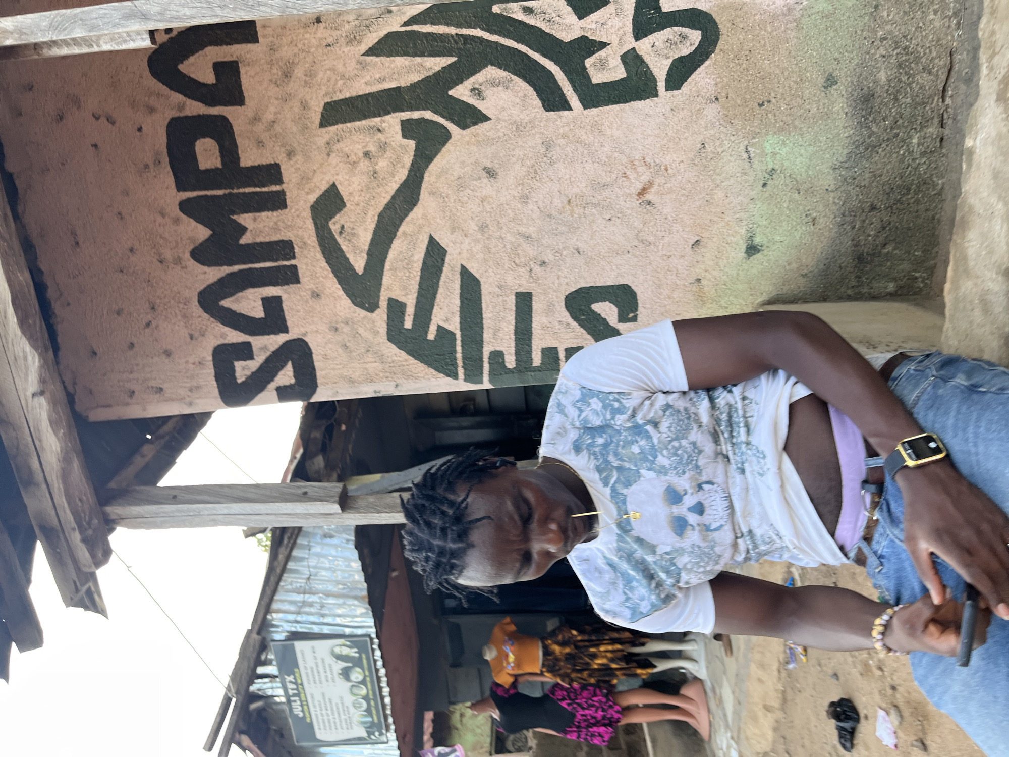 This image captures an outdoor scene, likely a street or market area, in Rafin Par, Nigeria. The setting features rustic structures with corrugated metal roofing and wooden supports.
The central figure is a young man with dark skin and dark braided hair. He wears a white t-shirt adorned with a blue and white patterned design featuring a skull graphic. He is dressed in blue jeans, revealing a light purple waistband beneath his shirt. A gold-colored chain with a small charm hangs around his neck. On his left wrist, he wears a black-strapped wristwatch with a gold-colored face and a beaded bracelet. He holds a mobile phone in his left hand, and his gaze is directed downwards, suggesting a moment of contemplation or interaction with his phone.
In the background, partially obscured, are other individuals and objects. One person is visible in an orange top, and another in a pink and black patterned dress stands near mannequins, hinting at a nearby retail or tailoring establishment.
A prominent feature in the upper part of the image is a light-colored wall with large, dark green graffiti. Clearly visible text includes "SAMPA" written vertically, and parts of other letters such as "STT" and an abstract symbol. In the lower-left background, a sign for a business is visible, bearing the text "JULI YX" along with unreadable smaller text and icons.
The lighting indicates it is daytime, with clear weather. The overall impression is a candid snapshot of daily life in an outdoor urban or semi-urban environment.