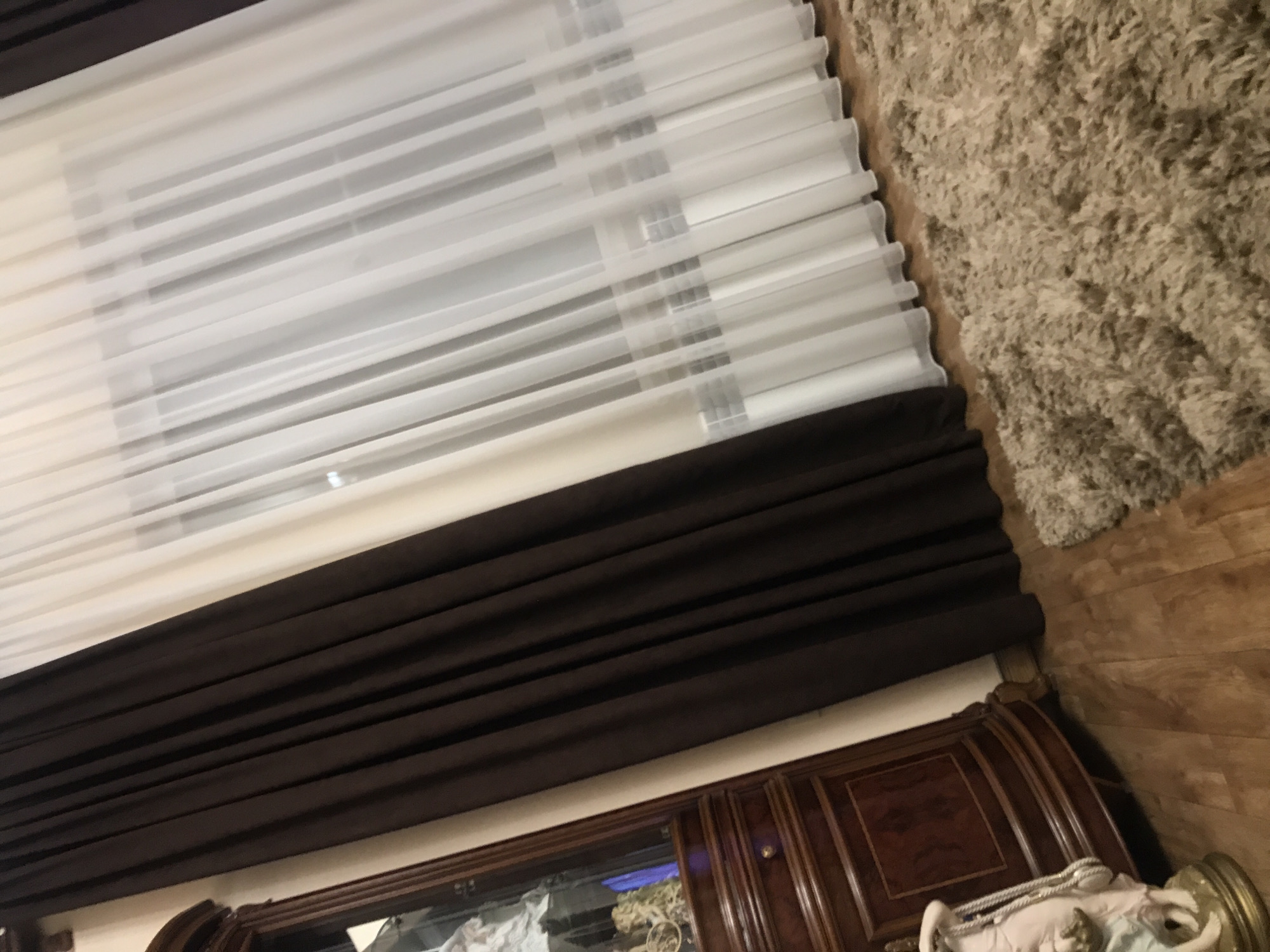 The image presents a low-angle, interior shot featuring a combination of window blinds, a heavy curtain, and a portion of an ornate wooden cabinet or headboard. The window blinds, rendered in a translucent white, occupy the upper left quadrant of the composition, running diagonally from the upper left to the center. Directly beneath the blinds is a dark brown curtain, gathered in heavy folds, obscuring most of the top of the wooden furniture. The top of the ornate cabinet is featured in the bottom portion of the image, showcasing wood paneling. The surface beside the curtains is partially covered with a shaggy rug. The location of the image is not discernible from the provided visual information, and therefore, the location in Yerevan, Armenia, cannot be determined.