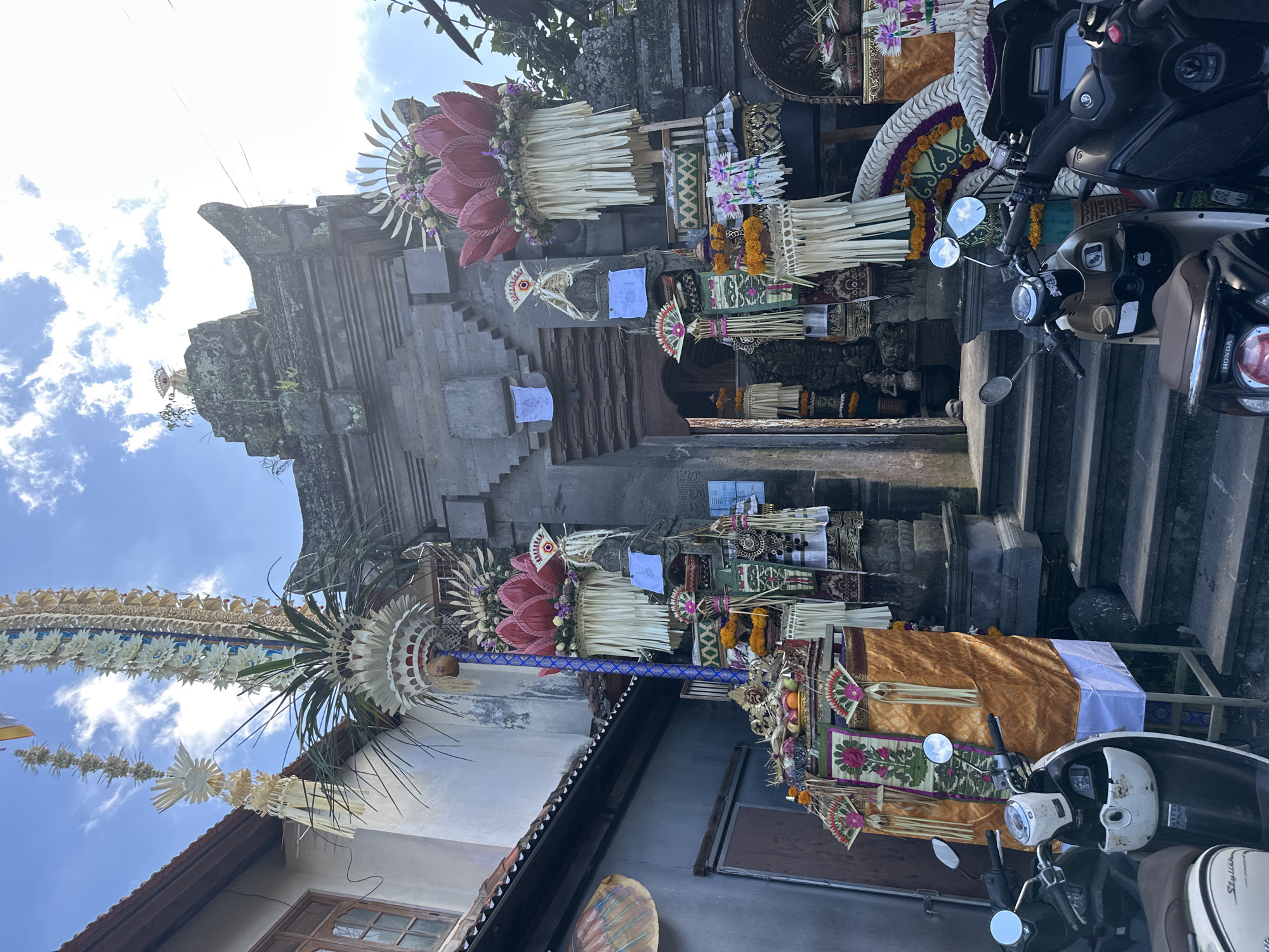 This image captures a vibrant street scene in the Ubud District of Indonesia, possibly during a religious festival or ceremony. In the foreground, several scooters are parked. The central focus is a traditional Balinese temple entrance, adorned with elaborate offerings and decorations. These include tall, ornate structures made of palm leaves, flowers, and woven materials, featuring large, lotus-shaped elements in pink and white. Smaller decorative fans and woven baskets filled with fruits and flowers are also visible. The temple gate itself is constructed of dark stone and appears weathered, with moss growing on its upper sections. To the left of the gate, a white building with wooden window frames and a tiled roof is visible, along with a tall ceremonial banner reaching towards the sky. The sky above is blue with scattered white clouds. The overall impression is one of rich cultural expression and community gathering in a tropical setting.