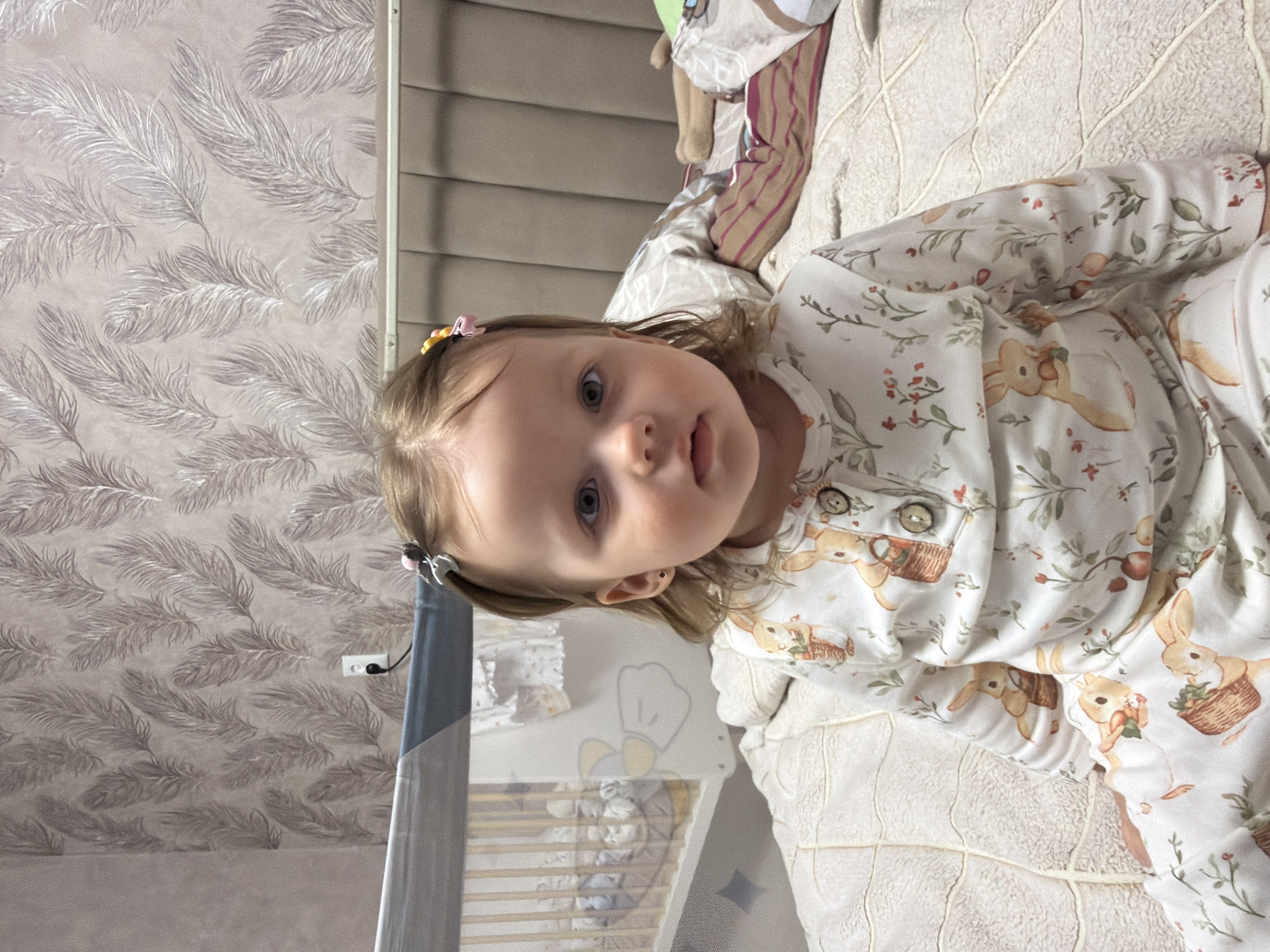 A young child with fair skin, light hair, and blue eyes is lying on a patterned blanket. The child is wearing a white pajama set adorned with illustrations of bunnies holding baskets of fruit. The background includes a wall with a feather-like patterned wallpaper, a partially visible cot with toys inside, and a section of window blinds. There is no evidence to determine the location in Reshetnikovo, Russia.