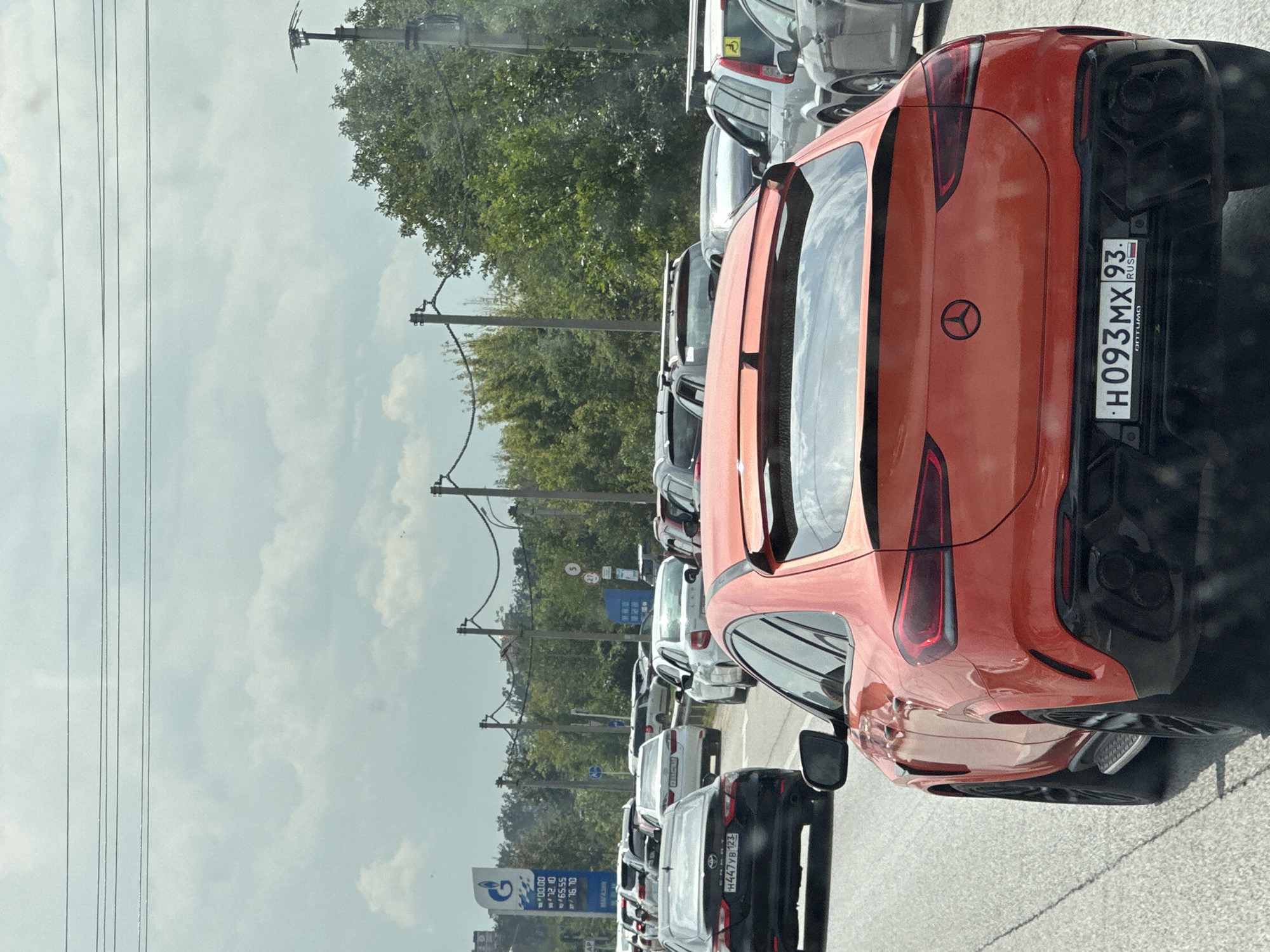 This is a medium shot, eye-level, horizontal, full-color photograph taken from inside a car on a cloudy day. The perspective is looking forward, capturing a traffic jam on a paved road. The foreground is dominated by the rear of a bright orange Mercedes-Benz GLE Coupe. Its license plate reads "H093MX 93 RUS". The car has a black spoiler and black accents around the taillights and bumper.
In the middle ground, several other cars are visible, including a black sedan with license plate "H447YB 123", a white sedan, and other vehicles in various colors. To the left of the road, a blue and white Gazprom gas station sign is visible. Overhead, power lines stretch across the sky. The background consists of trees lining the road and a hazy, overcast sky. The environment suggests a suburban or roadside setting within the city of Beryozovy, Russia, indicated by the gas station branding and license plate format.