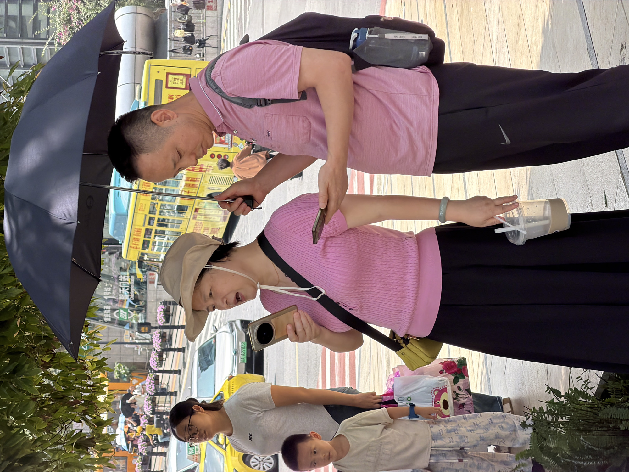 In Yuzhong District, China, on a sunny day, a group of people stand on a sidewalk near a street with a yellow bus and several cars. A man and a woman stand together under a large black umbrella. The man, wearing a pink polo shirt and black pants with a Nike logo, holds a smartphone and the umbrella. The woman, in a pink knitted shirt, a beige hat, and a black skirt, is looking at her phone and holding a drink. To their left, a young boy in a light-colored t-shirt and patterned pants stands looking forward. A woman in a grey t-shirt and glasses is also visible behind the boy, and other pedestrians and vehicles are in the background. The scene suggests a typical urban environment with people going about their day.