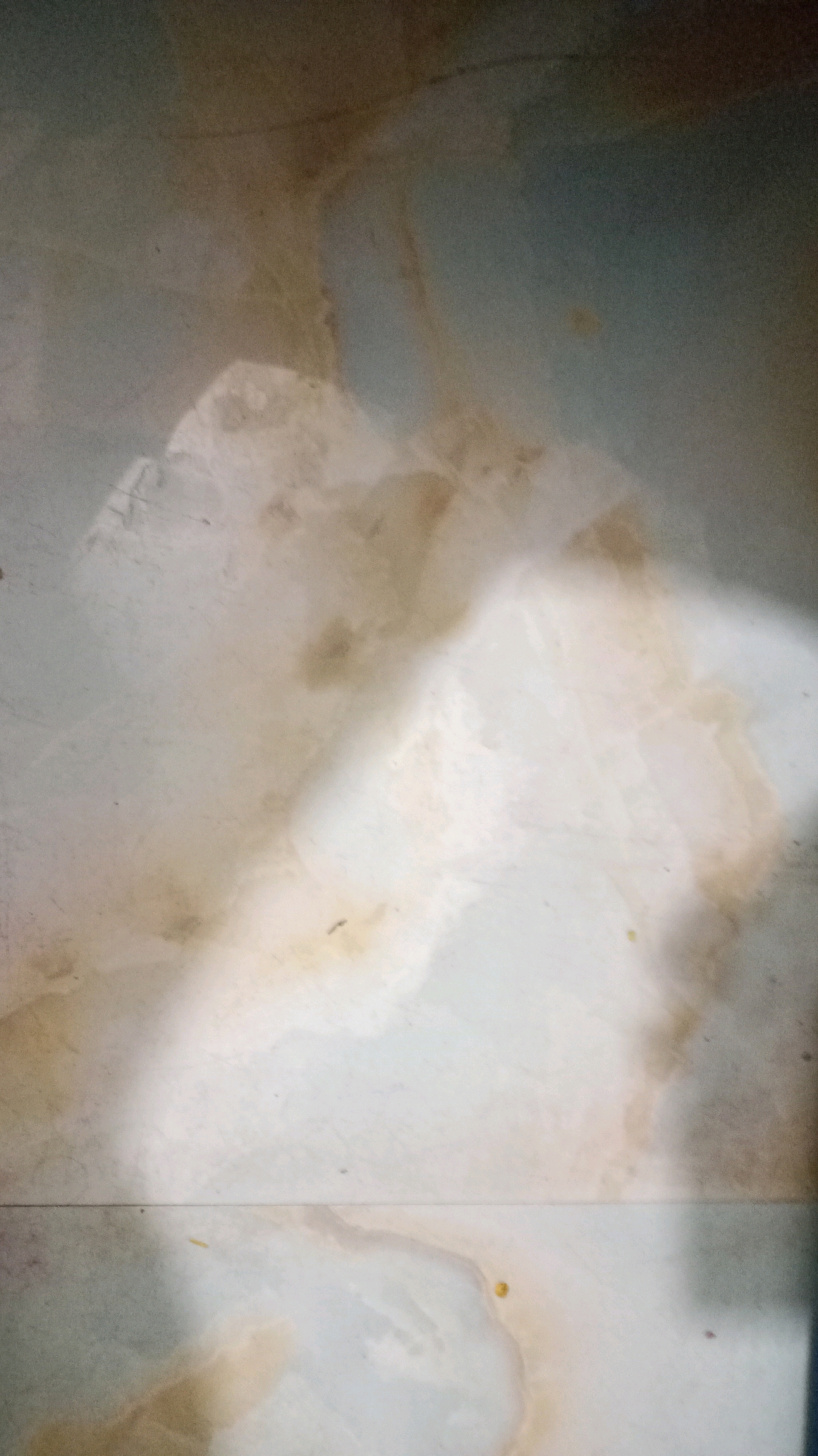 The image shows a close-up view of a tiled floor, likely in Deoghar, India. The tiles are light-colored with prominent brown and beige staining or marbling patterns. The lighting creates a strong diagonal shadow across the middle of the image, highlighting the textured surface of the tiles. There are a few small, dark specks scattered across the tiles, which could be dirt or debris. The overall impression is of an aged or heavily used floor.
