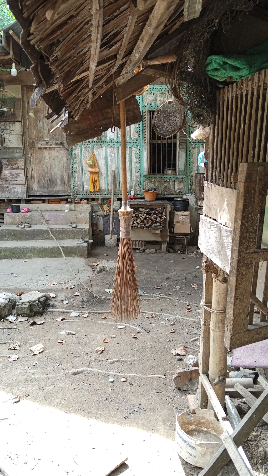 This image captures a scene in the city of Tanjong Krueng, Indonesia. In the foreground, a broom made of natural fibers hangs from a wooden pole. The broom appears well-used, with its bristles spread out. Behind the broom, there are worn steps leading up to a rustic wooden building. The building's facade is weathered, with visible planks and a wooden door. A window with bars is visible on the building. The ground is a mix of dirt, small stones, and scattered dry leaves. Various objects are scattered around the area, including a bucket, some discarded items, and what appears to be firewood stacked near the building. The overall environment suggests a rural or impoverished setting, with a focus on functional, everyday items and a lack of modern amenities.