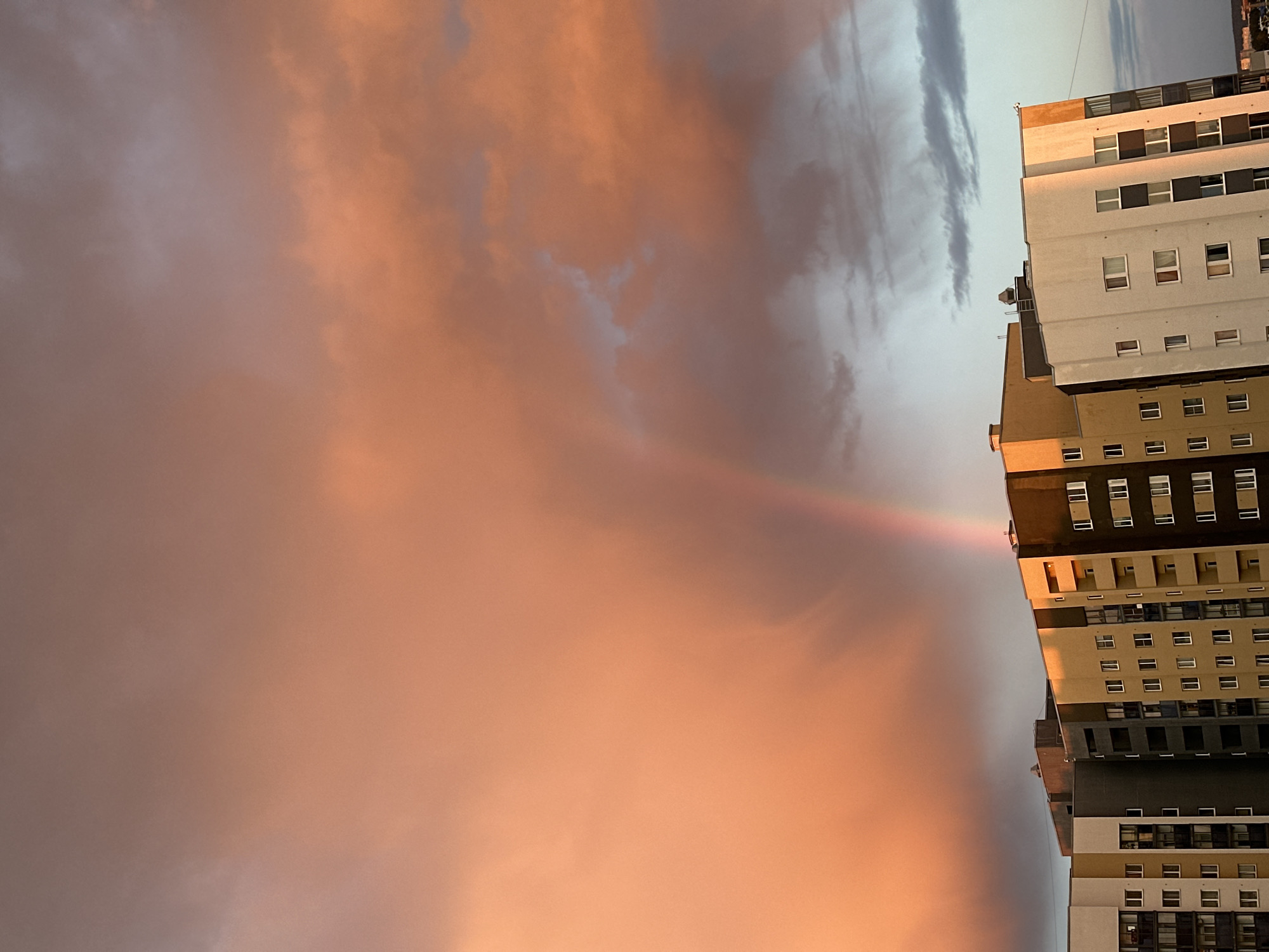 The image captures a building with a rainbow appearing to emanate from it. The building is multi-story with a visible grid of windows and varying colors on different sections, ranging from white to beige to dark grey. The sky is filled with peach-colored clouds, likely from a sunset or sunrise. The rainbow extends from the building into these clouds. The overall perspective is a tilted upward view of the building against the dramatic sky. The location is Yekaterinburg, Russia.