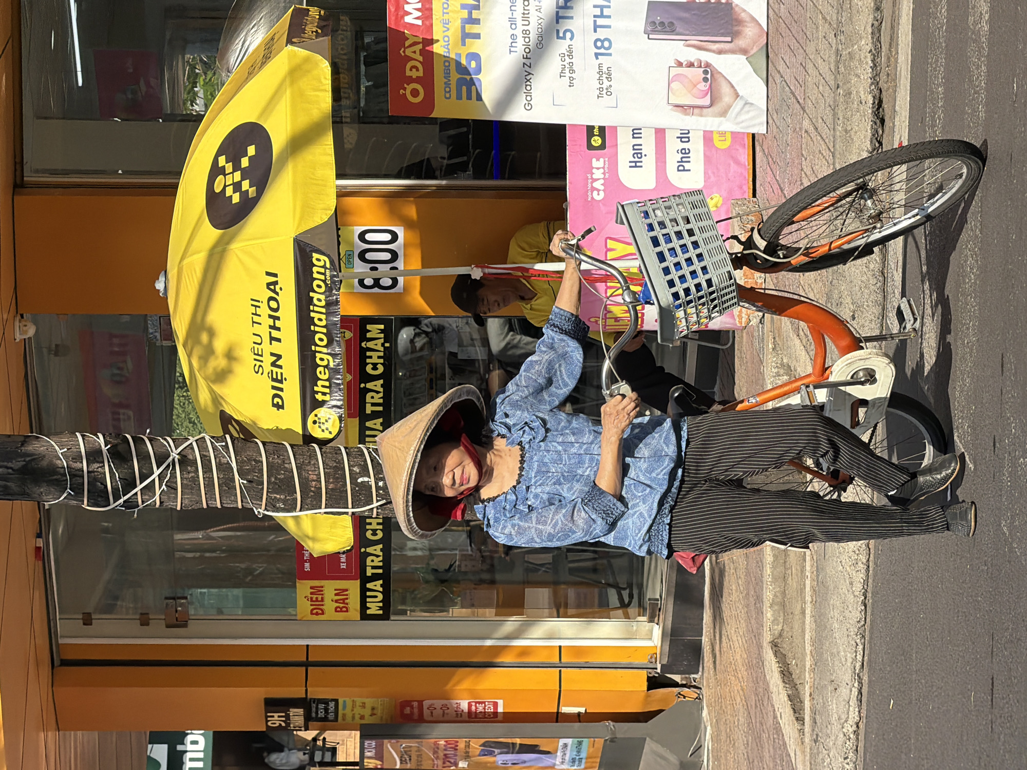 A Vietnamese woman wearing a conical hat, a blue patterned shirt, and striped pants stands next to an orange bicycle with a basket on the street in Nha Trang, Vietnam. She is looking to her right. A yellow umbrella with Vietnamese text and a logo is open above her. In the background, a store with an orange facade displays advertisements. A man wearing a yellow shirt and a dark cap is visible inside the store. The scene appears to be a street-level commercial area with pedestrian traffic.
