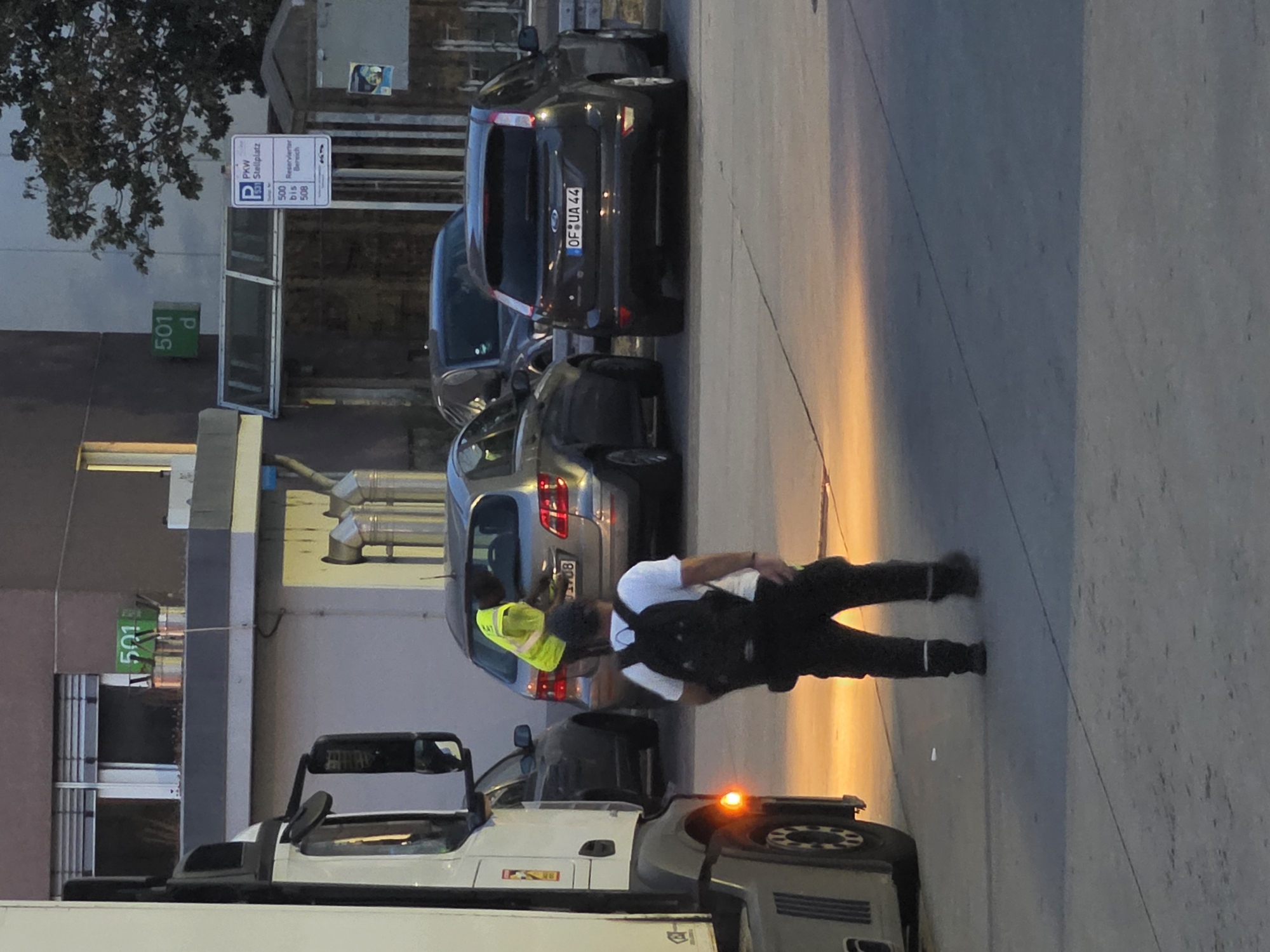 A person wearing a high-visibility vest and a backpack is standing in a parking lot in Frankfurt, Germany. They are facing away from the camera, and the backpack is black. A truck is parked to the left of the frame. A gray car is parked in the background. A parking sign is visible in the background with the text "PKW Stellplatz" and "Reservierter Bereich". There are also two green signs on a building in the background with the numbers "501" and "501 d". The ground is paved and appears to be asphalt. The lighting suggests it is either early morning or late evening.