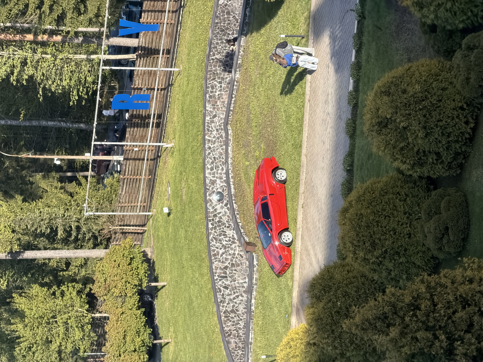 This image shows an aerial view of a park-like area in Repino, Russia. A red sports car is parked on the grass next to a stone pathway. Two people stand near the walkway, appearing to be taking a picture. In the background, a wooden structure with blue letters spells out "REPINO." Trees surround the area.