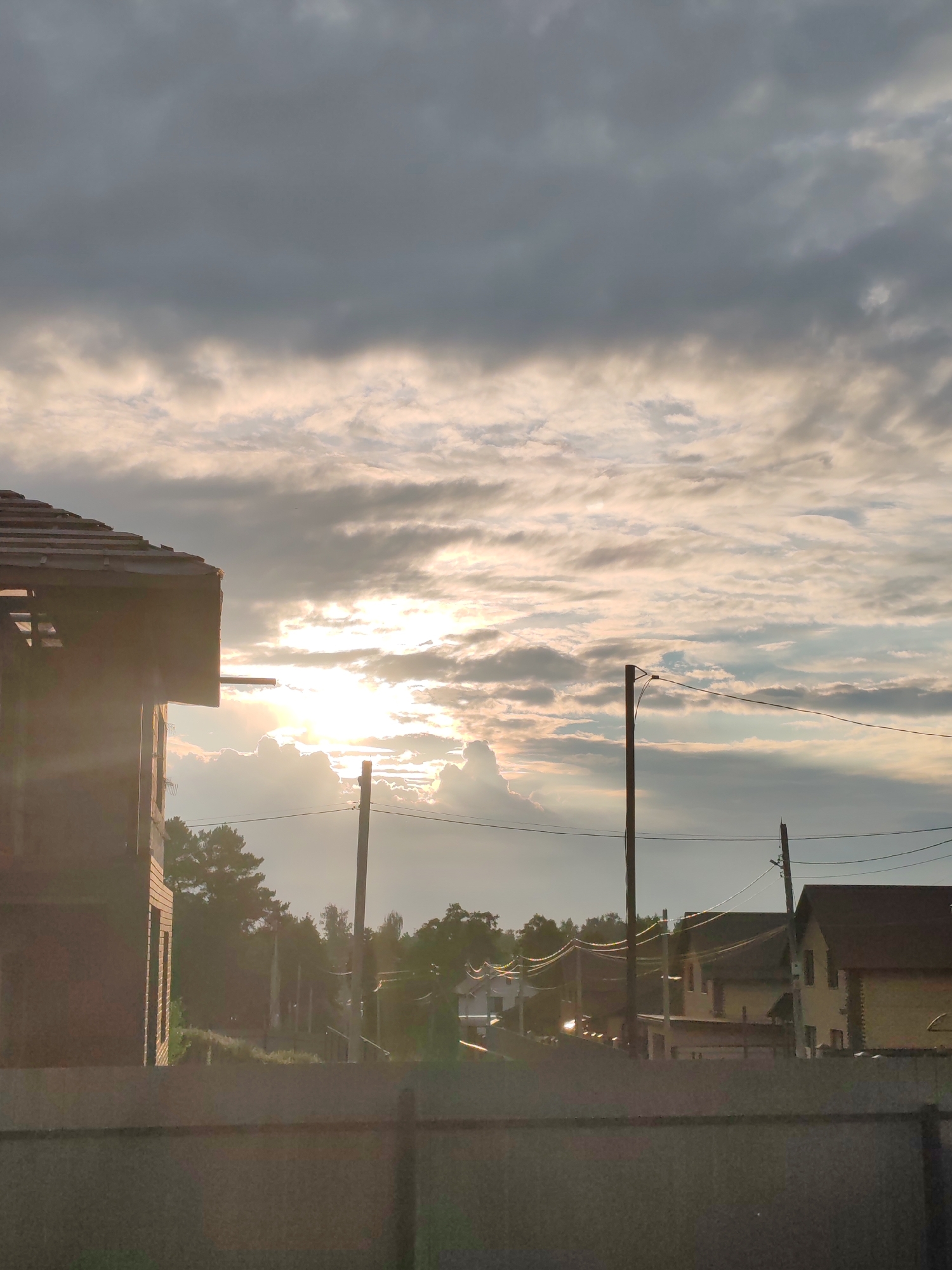 This media file depicts an outdoor scene with a cloudy sky as the primary subject. The clouds are varied in texture and color, with patches of bright sunlight breaking through, suggesting a dynamic weather condition.
In the foreground, a large, unfinished brick building with exposed wooden roof beams is visible on the left side, partially obscuring the view. Behind it, a line of trees and several houses can be seen, indicating a residential or semi-rural setting. Power lines with wooden poles stretch across the frame, adding to the sense of a developed area. The lighting suggests it is either sunrise or sunset, with the sun's rays casting a warm glow and creating lens flares.
The overall mood is serene, with the dramatic sky providing a backdrop to the ordinary elements of the landscape. No people are visible in the image. No text is present. The scene is likely located in Chaikovsky, Russia, given the contextual information.