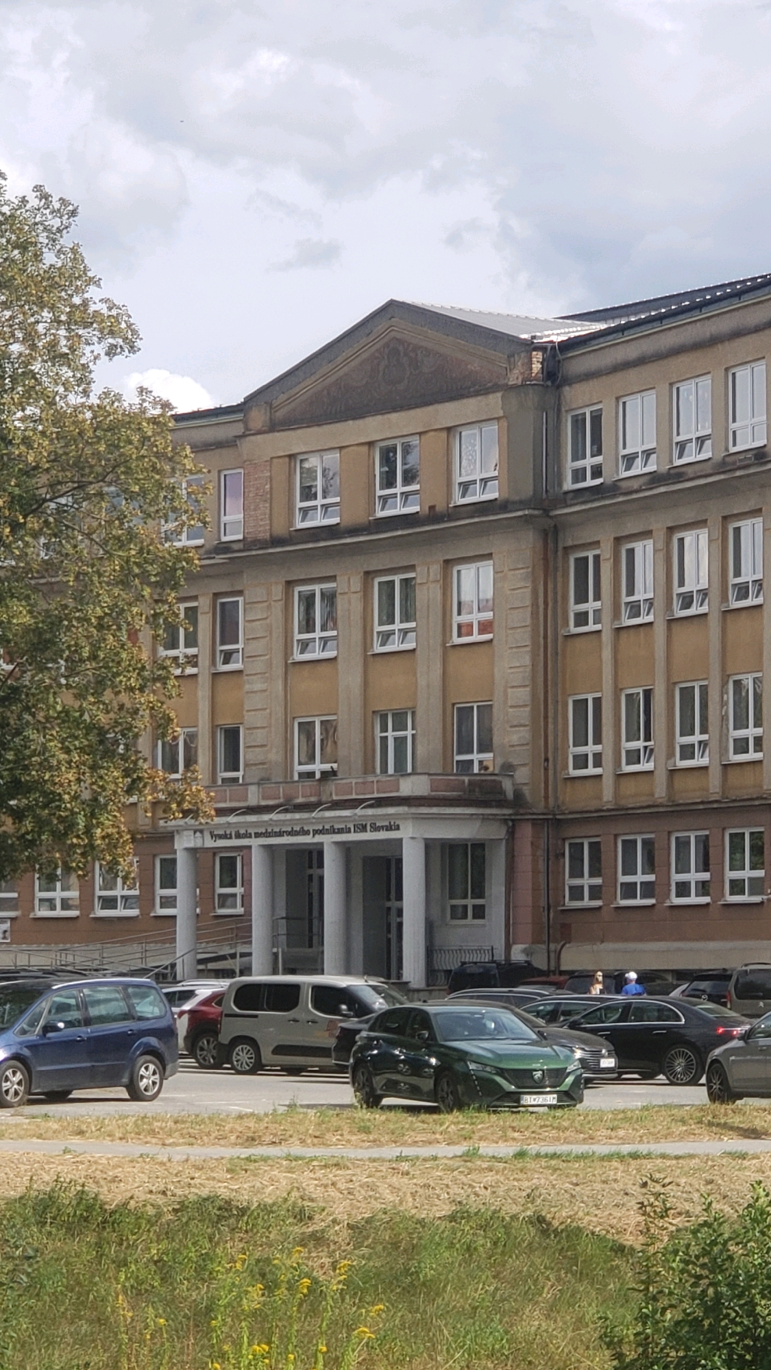 The image shows the facade of a multi-story building with a beige exterior and numerous windows. Above the entrance, there is a sign that reads "Vysoká škola medzinárodného podnikania ISM Slovakia". The building appears to be an educational institution. In front of the building, there is a parking lot with several cars parked, including a blue minivan, a silver van, a green sedan, and other vehicles. The area is partially covered with grass and dry vegetation, suggesting an outdoor setting. The sky is partly cloudy. This building is located in Prešov, Slovakia, and is identified as the International School of Business and Management (Vysoká škola medzinárodného podnikania ISM Slovakia) in Prešov.