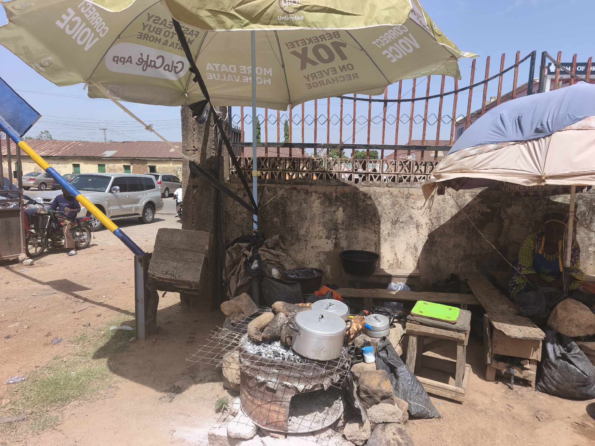 This media file depicts an outdoor food stall or market scene in Jos, Nigeria, under bright, clear daylight, casting strong shadows. The central focus is a rustic cooking setup: a metal barrel charcoal grill, supported by rocks, on which two aluminum pots and what appears to be roasted yams or plantains are being heated. Wooden stools, planks, a green cutting board, a small white container, a black bowl, and several black plastic bags with goods are arranged around the cooking area, indicating an active commercial space.
Overhead, two large umbrellas provide shade. The primary one, a light yellow-beige, displays mirrored text from a mobile network provider, including "GLO Unlimited," "CORRECT VOICE," "XOX BEKEKELEI," "ON EVERY RECHARGE," and "GLO Café app." A second, blue and beige patchwork umbrella is partially visible to the right.
In the mid-ground, a blue and yellow striped barrier pole separates the stall from a street with light traffic, including a silver SUV and a person on a motorcycle. Further in the background, a metal fence and buildings with rusty tin roofs complete the scene, characteristic of a bustling roadside or market environment. A faint sign in the background appears to read "STOP DISCRIMINATING" or "STOP ORINATING." The image captures a moment of daily life and commerce, with an emphasis on local food preparation.