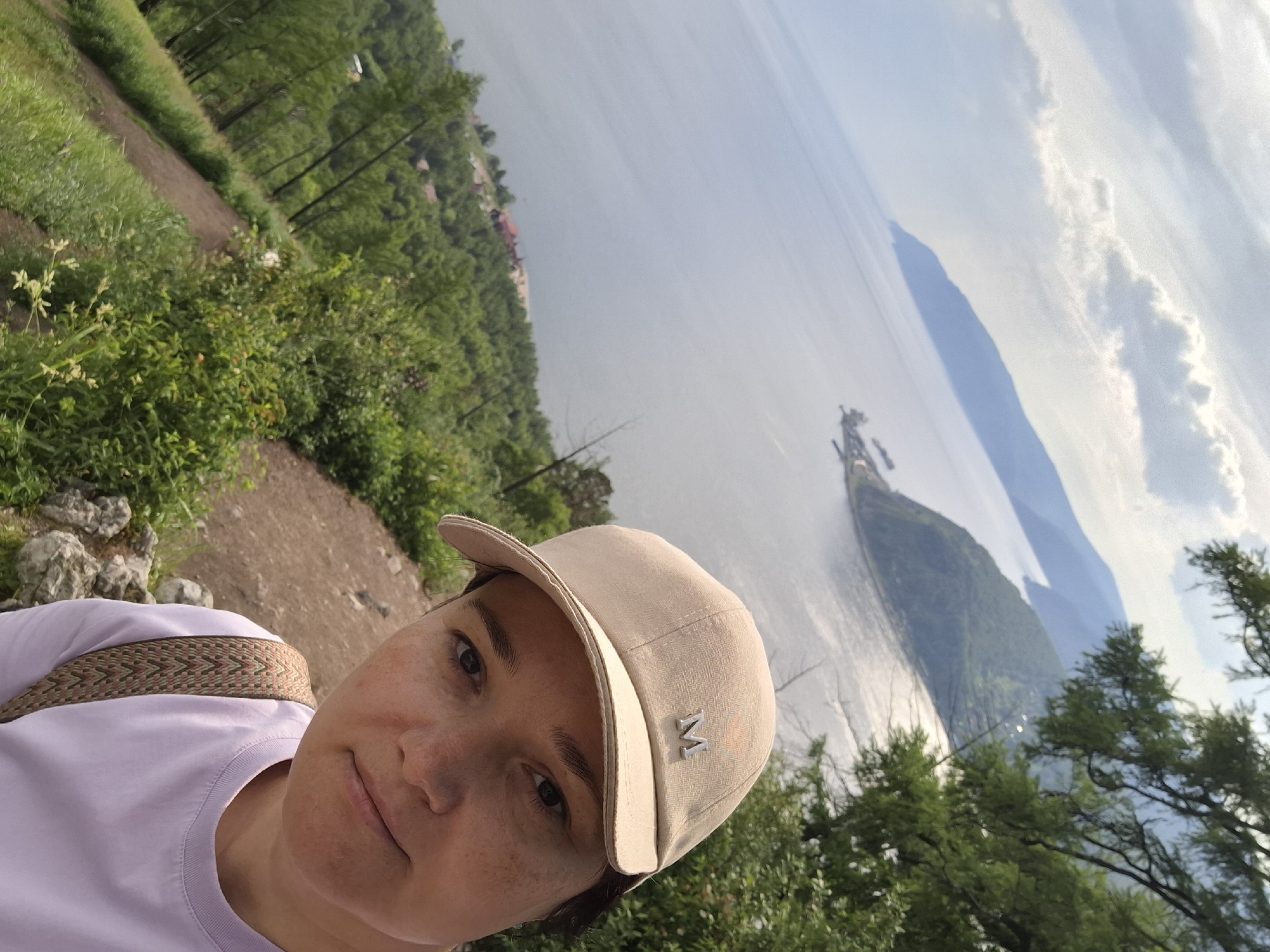 This media file is a selfie taken by a person wearing a light purple t-shirt and a light beige baseball cap. The cap has a silver emblem that appears to be the letter "M". The person has fair skin with freckles and dark eyes. They are smiling slightly at the camera.
The background of the image shows a scenic view of a large body of water, likely a lake, with mountains in the distance. The water is calm with reflections of the sky and surrounding landscape. There are green trees and vegetation on the left side of the image, with a dirt path winding through the foliage. The lighting suggests it is daytime, possibly late afternoon, with a bright sky and some clouds. The overall mood appears relaxed and outdoorsy.
The setting is likely a natural landscape, possibly a hiking trail or viewpoint overlooking a lake. Given the location context of Nikola, Russia, this could be a view of a lake in that region. There are no other people visible in the image.