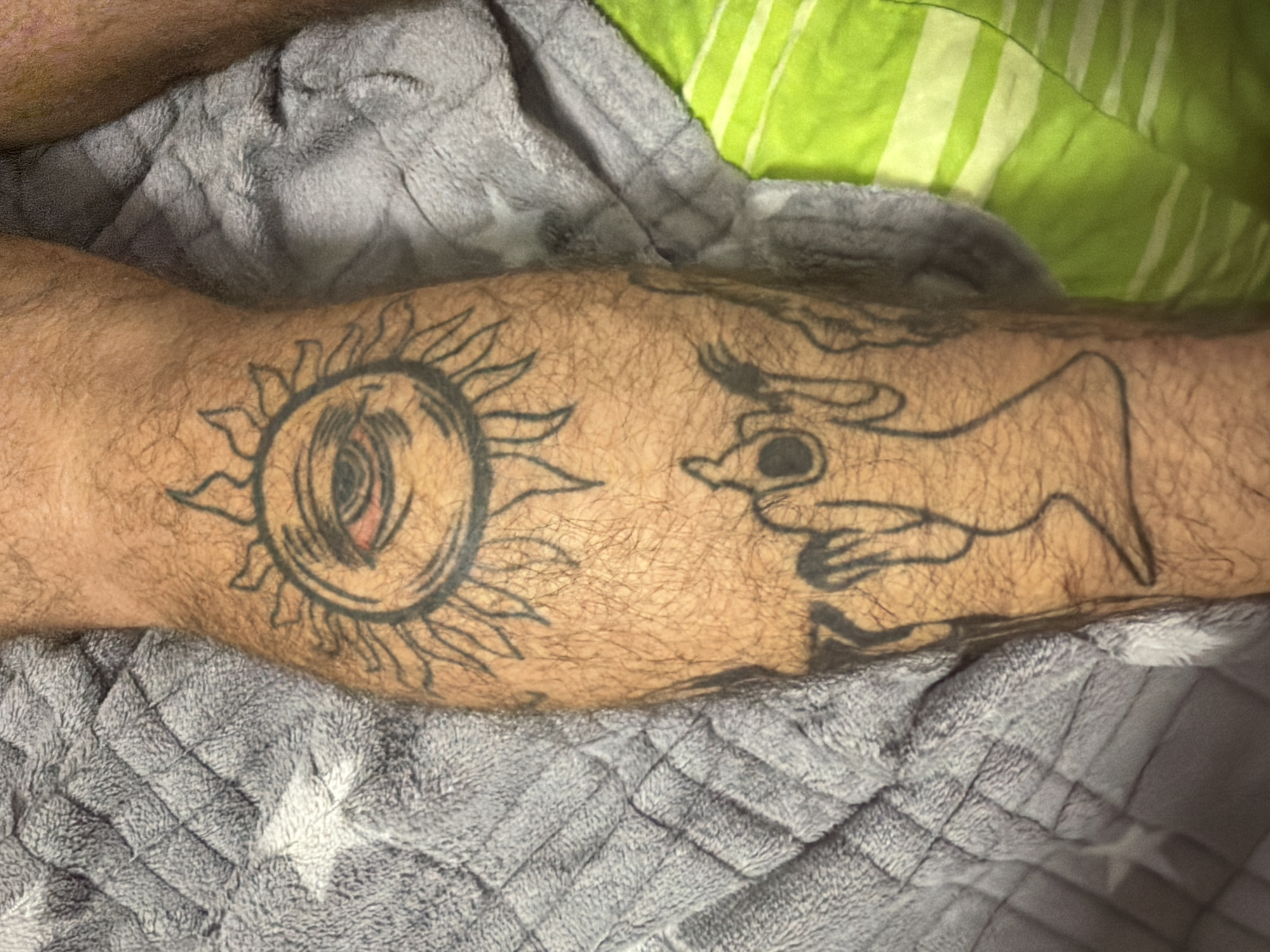 The image displays a tattoo on a person's leg, featuring a sun with an eye in the center, and a ghostly figure below it. The leg is covered in hair and is resting on a gray, textured blanket with a star pattern. To the right of the leg, a bright green blanket with white stripes is visible. The tattoo is the primary subject, with the eye in the sun appearing red and possibly irritated. The background suggests an indoor setting, possibly a bed or sofa, given the blankets. No specific location in Dorsten, Germany can be determined from the visual information provided.