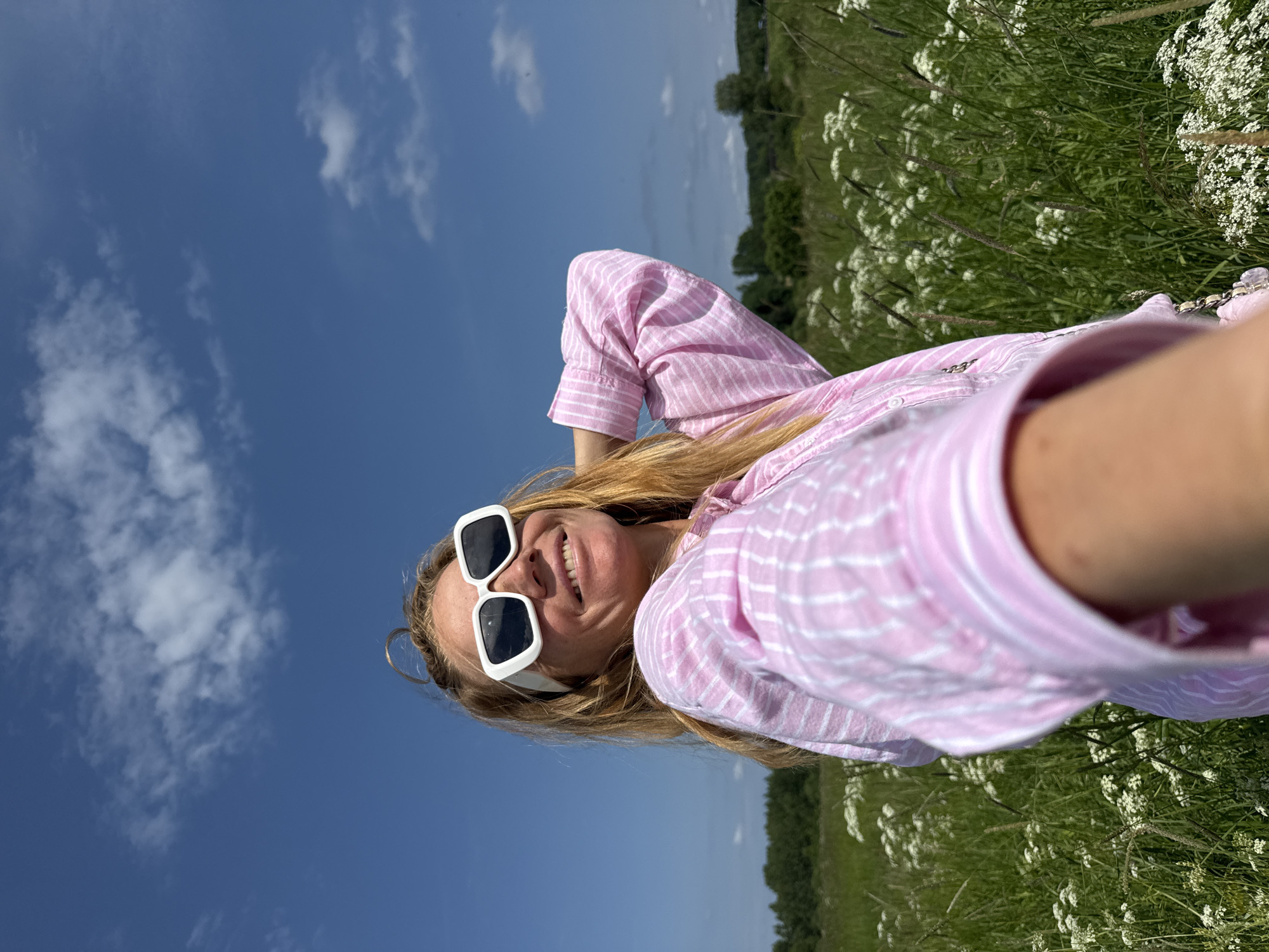 A woman with blonde hair and white-rimmed sunglasses is taking a selfie in a grassy field. She is wearing a pink and white striped button-down shirt and smiling. The background shows a clear blue sky with some scattered clouds, green grass, and small white flowers. The location is not specified as Menevsha, Russia, or any other city.