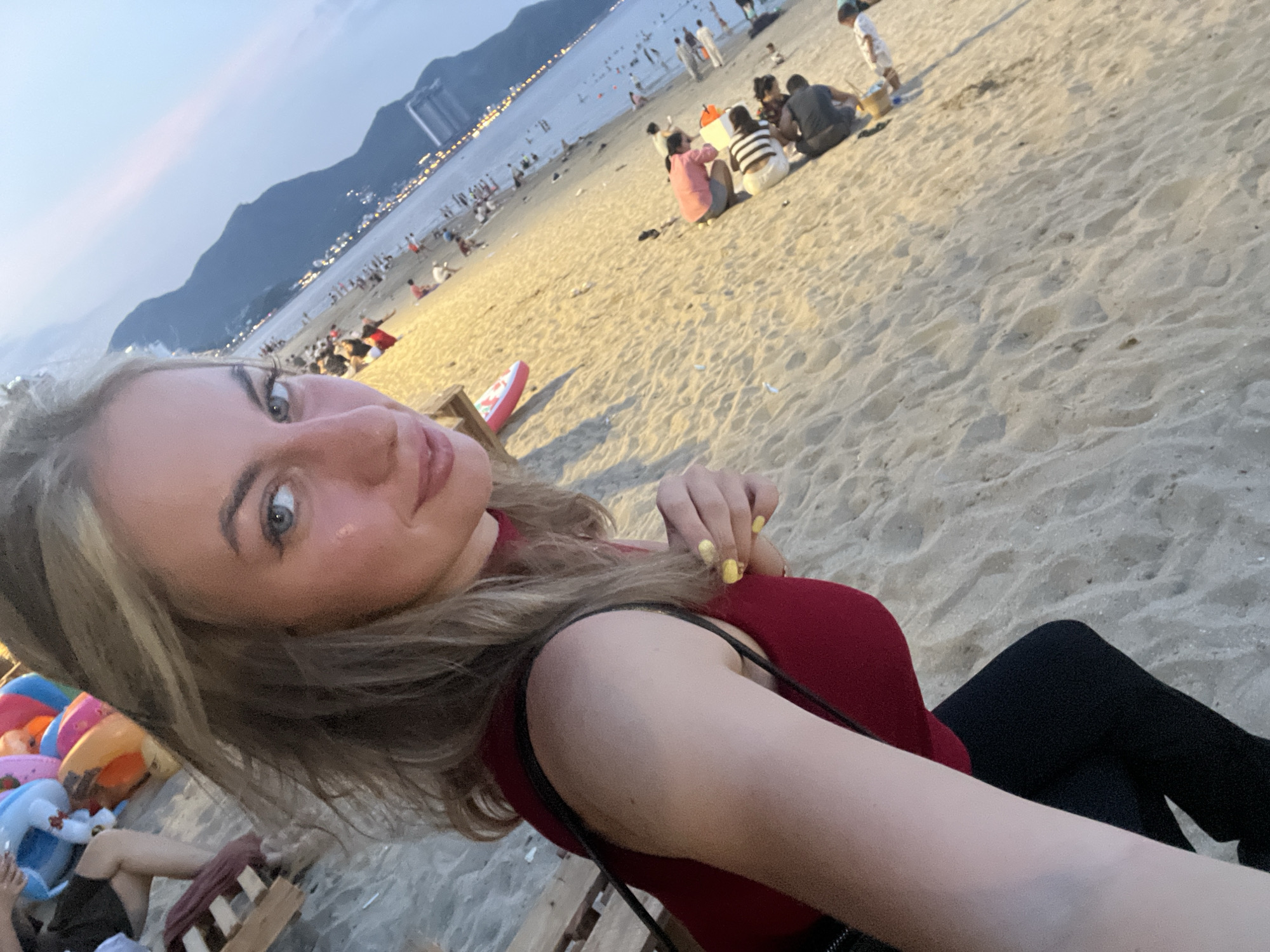An outdoor, eye-level, medium shot shows a young woman with blonde hair taking a selfie on a sandy beach. The woman is wearing a red top and black pants, and her nails are painted yellow. She is smiling at the camera. In the background, other people are visible on the beach, some sitting and some standing. The beach appears to be crowded, with some beach umbrellas and other beach equipment scattered around. The water is visible in the distance, and mountains can be seen on the horizon. The sky is a pale blue, and it appears to be either early morning or late evening. The overall mood of the image is relaxed and casual.