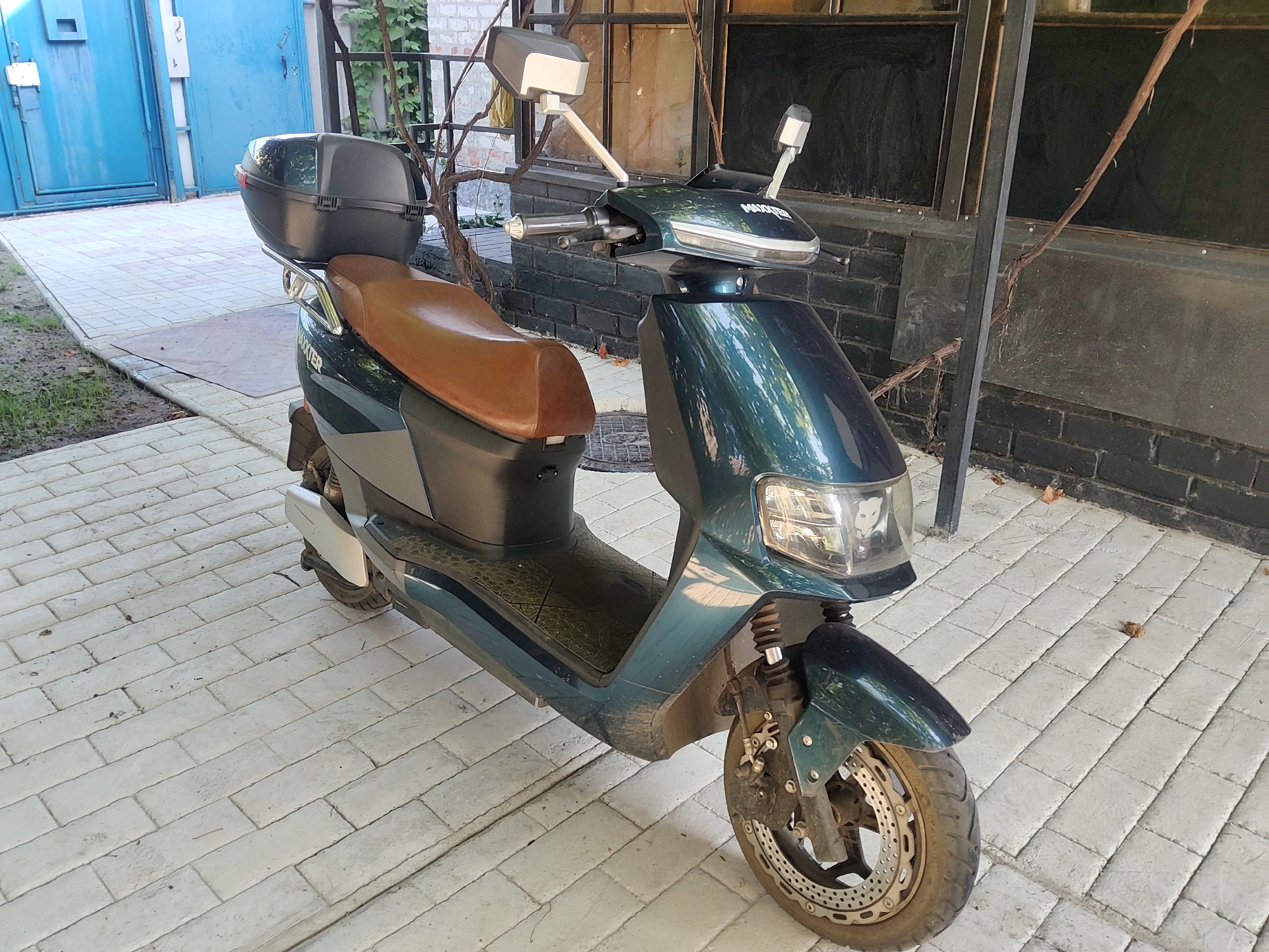 A teal-colored electric scooter is parked on a light-colored paved walkway. The scooter has a brown seat, a black storage box mounted on the rear, and a silver-colored front fender. The scooter is facing slightly to the left. In the background, there is a blue metal gate and a brick wall. The scene appears to be outdoors, possibly in a residential area or a courtyard in Kharkiv, Ukraine.