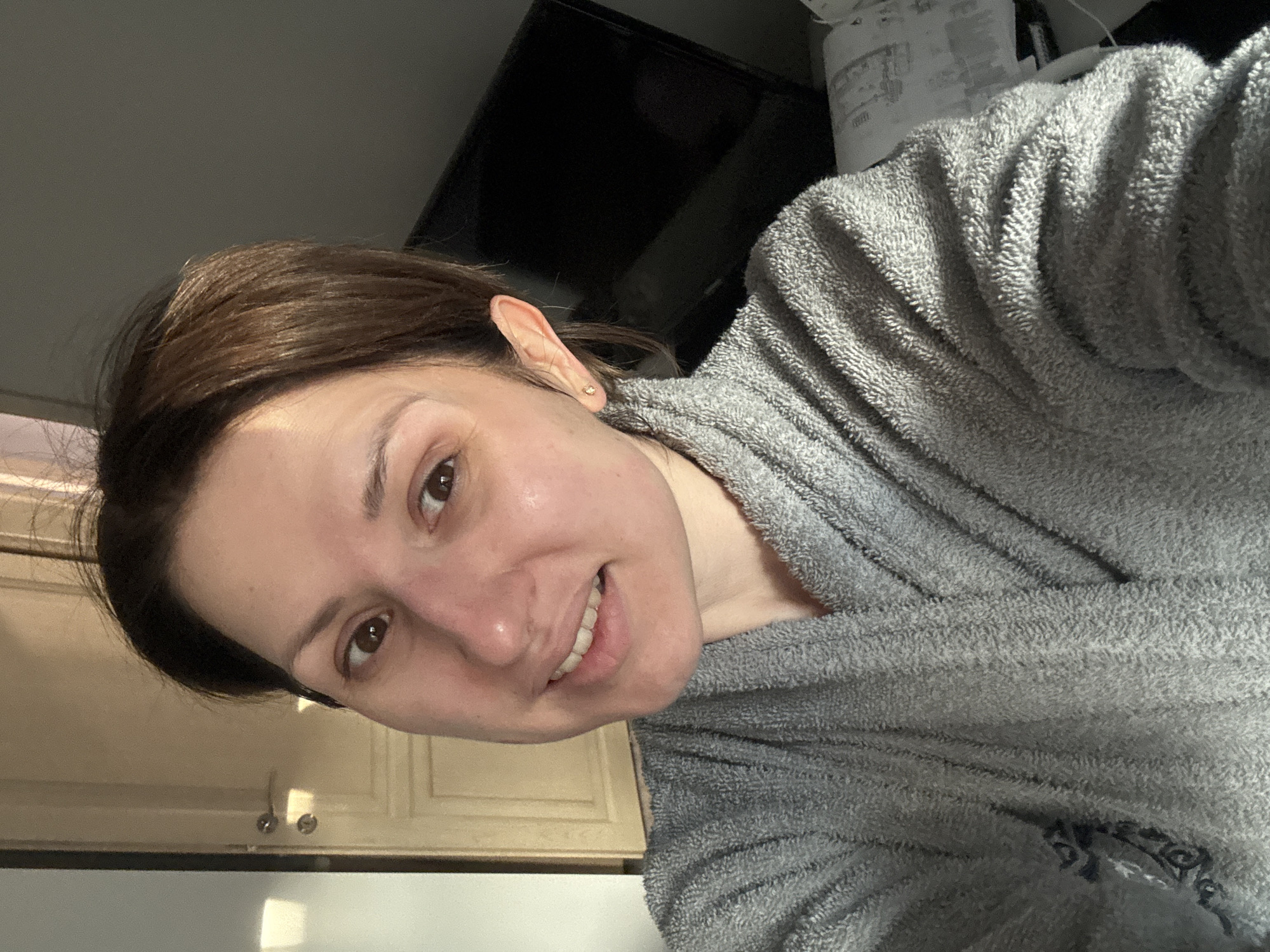 A woman with brown hair and brown eyes, wearing a gray robe, is in what appears to be a kitchen setting. Cabinets are visible in the background. A black television is partially visible in the upper background. There is no information in the image to determine if she is in Sofia, Bulgaria.