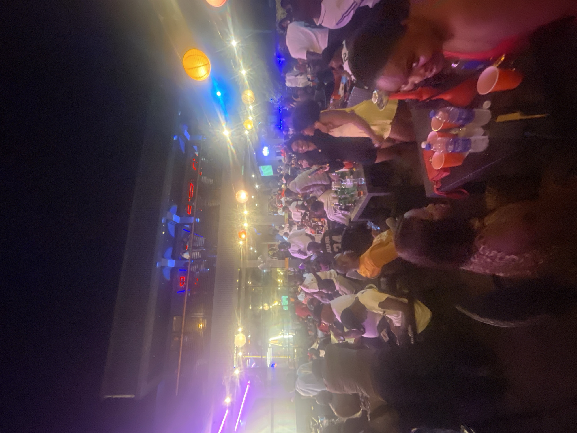The image shows a crowded outdoor event at night, possibly a club or bar, in Lokoja, Nigeria. People are seated at tables, drinking and socializing. There are numerous lit-up decorations, including hanging lights. There are people of various ages and genders. Tables have beverages, including water bottles and plastic cups.