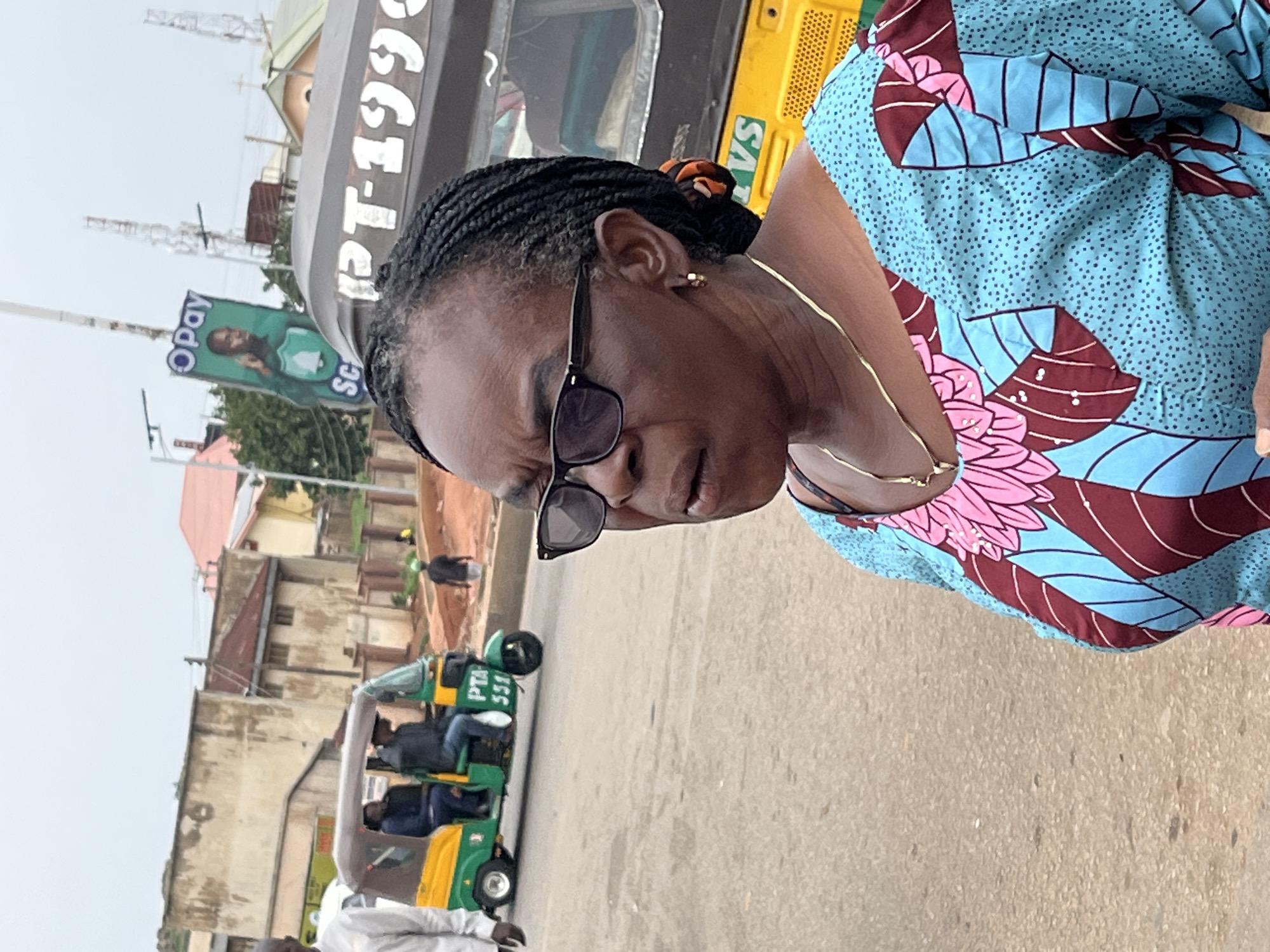 The image shows a woman wearing glasses and a colorful patterned dress, standing on a street. Behind her, a green and yellow tricycle, known as an "okada" or "keke napep," is visible, with passengers inside. There is also an advertisement for "Opay" with a picture of a man. The background shows buildings and utility poles, indicative of an urban environment. The scene appears to be outdoors on a cloudy day in Gura Top, Nigeria.