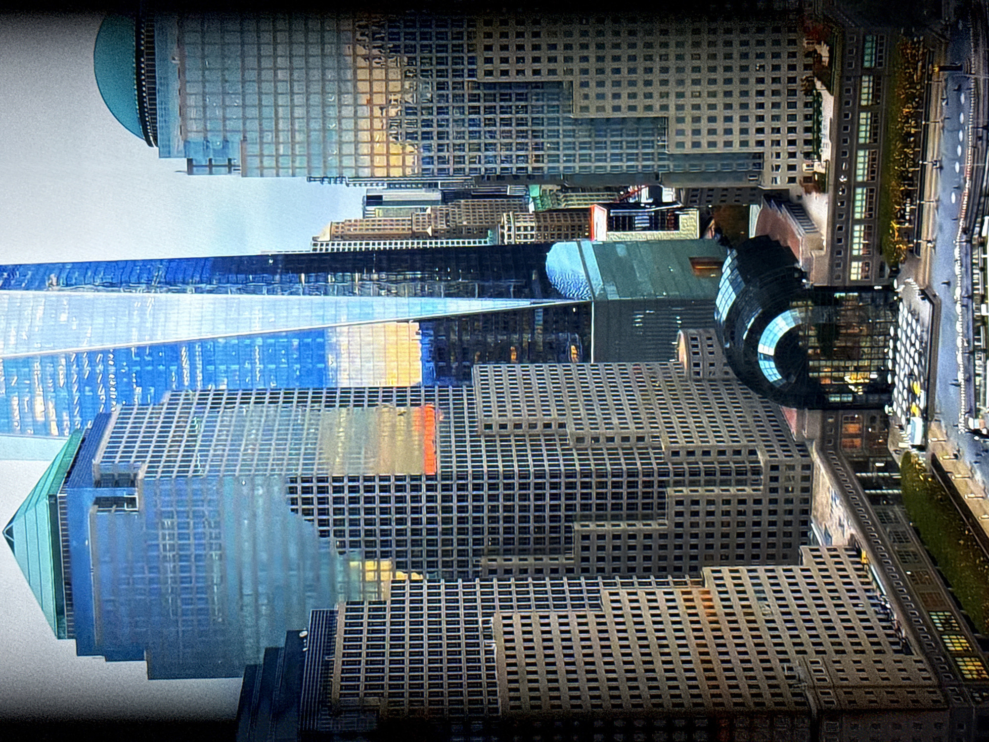 The image shows a collection of tall skyscrapers in a dense urban environment. The buildings are predominantly modern, constructed with glass and steel, reflecting the sky and surrounding structures. One prominent skyscraper in the center has a sharp, angled design and is significantly taller than the others. To the right, another building features a distinctive turquoise dome. The foreground shows a lower-level structure with a rounded, glass-enclosed roof, possibly a public space or transportation hub. Below this, there is a plaza area with trees, streetlights, and what appear to be pedestrian walkways and possibly some vehicles. The overall impression is of a bustling city center, possibly at dusk or on an overcast day given the lighting conditions.
This image does not depict the city of Burgas, Bulgaria. The architectural style and the specific buildings visible are characteristic of major financial districts in North American cities, particularly New York City, with the prominent skyscraper in the center strongly resembling One World Trade Center.