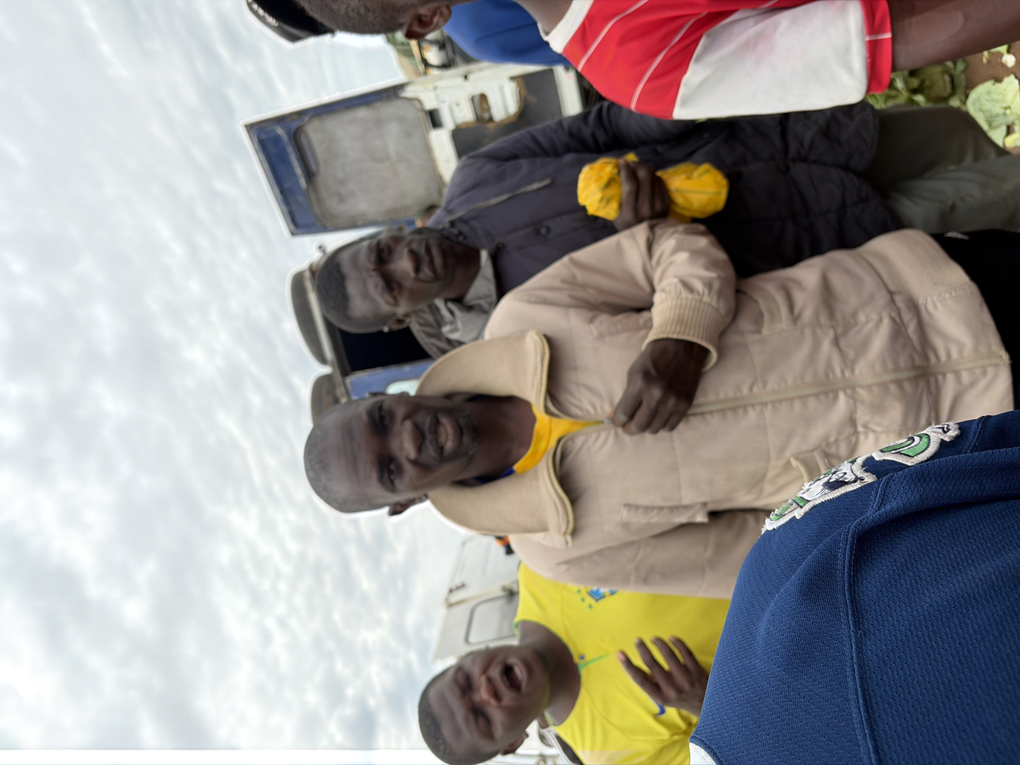 In Gura Top, Nigeria, under a cloudy sky, a group of men stand near a large vehicle with its door open. A man in a tan jacket with a light-colored collar is in the foreground, facing forward with his mouth slightly open. To his left, another man in a bright yellow shirt with a crest is visible, his mouth open and his hands gesturing. To the right of the man in the tan jacket, a man in a dark jacket holds a crumpled yellow cloth. Further to the right, a man in a red and white striped shirt is partially visible. The ground appears to be dirt, with some green leafy vegetables scattered around. The overall impression is a gathering of people, possibly in a market or at a roadside stop.