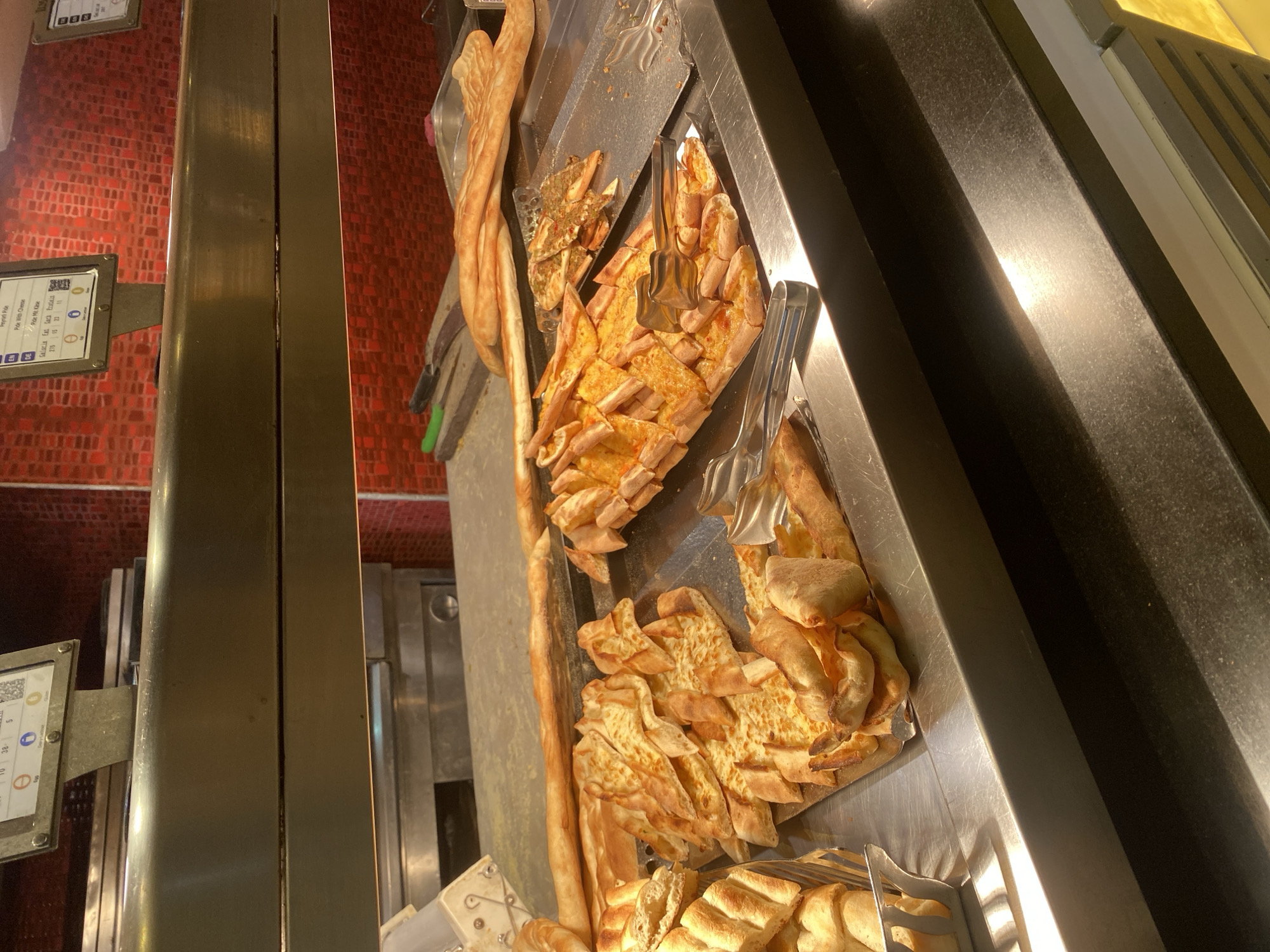 The image depicts a food service counter, likely in a restaurant or buffet, featuring a variety of Turkish-style flatbreads. The primary focus is on several trays of "pide," a boat-shaped Turkish pizza, some with cheese and others with meat toppings. In the foreground, there are also loaves of long, thin bread. A metal basket to the left holds more bread. Serving tongs are present on the trays. Behind the food, a stainless steel counter and a red tiled backsplash are visible. Above the counter, a metal shelf holds informational displays with text and icons. The overall environment appears to be clean and well-lit, suggestive of a food establishment. Without further context or specific landmarks, it is impossible to definitively place this scene in Aksu, Turkey.