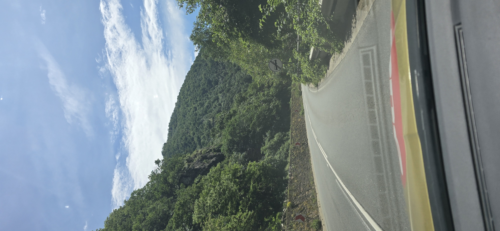 This image depicts a scenic mountain road captured from inside a vehicle on a bright, sunny day. The dominant feature is a steep, heavily forested mountain slope on the left, covered in lush green deciduous trees, with some rocky sections visible through the foliage. Above the mountain, the sky is a clear, vibrant blue with scattered, wispy white clouds.
A paved asphalt road with a distinct double white centerline winds gently to the right, following the natural curvature of the terrain. Along the left edge of the road, a stone retaining wall is visible, punctuated by several red and white reflective hazard markers, likely indicating the road's curve. Further up the slope, partially obscured by tree branches, a circular white road sign with a black symbol and a red diagonal line suggests a traffic restriction, such as "no overtaking" or "no entry for specific vehicles." A metal guardrail is also faintly visible behind the trees.
The rightmost edge of the photograph includes elements of the vehicle's interior, specifically a yellow strip and a dark gray or black panel, indicating the image was taken from a car or bus window. The bright sunlight suggests it is midday, and the weather appears clear and pleasant, ideal for a journey through this mountainous landscape. No explicit text is readable, only the symbolic road sign.