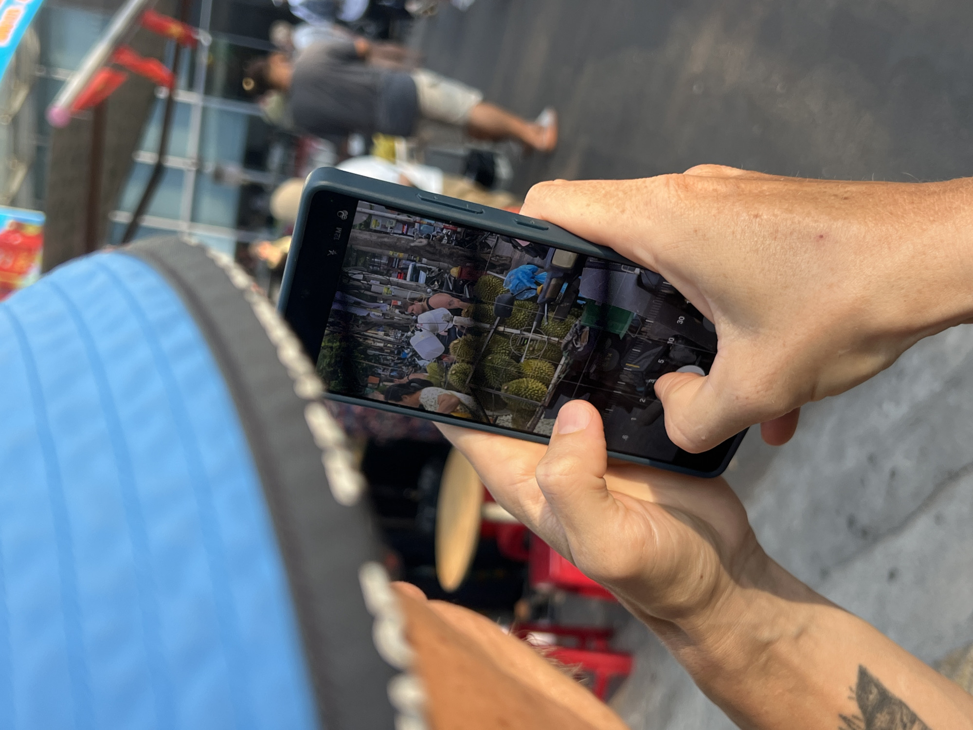 A person is taking a photograph with a smartphone of a scene in Nha Trang, Vietnam. The smartphone's screen shows a fruit stand selling durian. Other people are present in the scene captured by the phone's camera. The person taking the photo is standing on a paved surface. A blue object, potentially a bag or part of clothing, is visible in the foreground near the bottom left corner.
