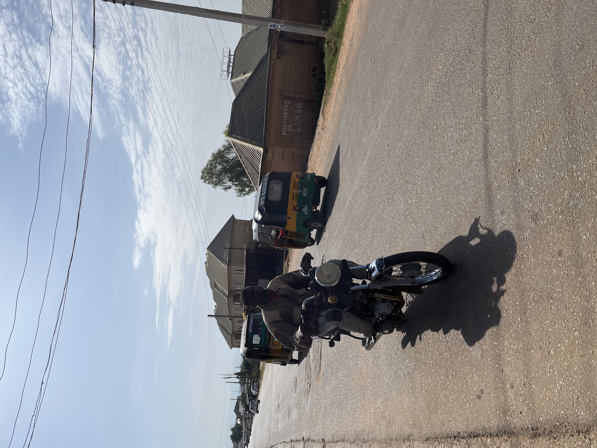 A man sits on a motorcycle on a paved road in Jos, Nigeria. A three-wheeled vehicle, sometimes called a rickshaw or keke, is parked nearby. Buildings line the street, including a brick structure with a corrugated metal roof. Overhead wires run along poles visible in the image. The sky is light blue with some clouds.