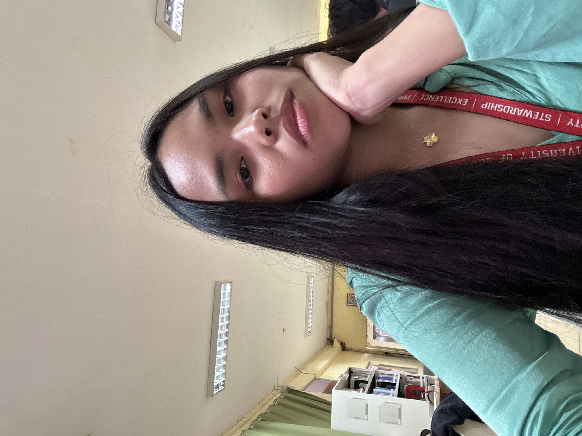 The image shows a person with long, dark hair, light skin, and facial features that suggest they are of Filipino descent. They are wearing a light green shirt or top. Around their neck is a red lanyard with the words "UNIVERSITY OF DAVAO" and "STEWARDSHIP | EXCELLENCE | PROFESSIONALISM" printed on it. They are touching their face with their hand.
The background appears to be an interior space, possibly a classroom or office, with fluorescent lights on the ceiling. There is office equipment or supplies in the background. The lanyard suggests a location within the University of Davao. The presence of the lanyard, clothing, and facial features strongly indicate the setting is in Davao City, Philippines.