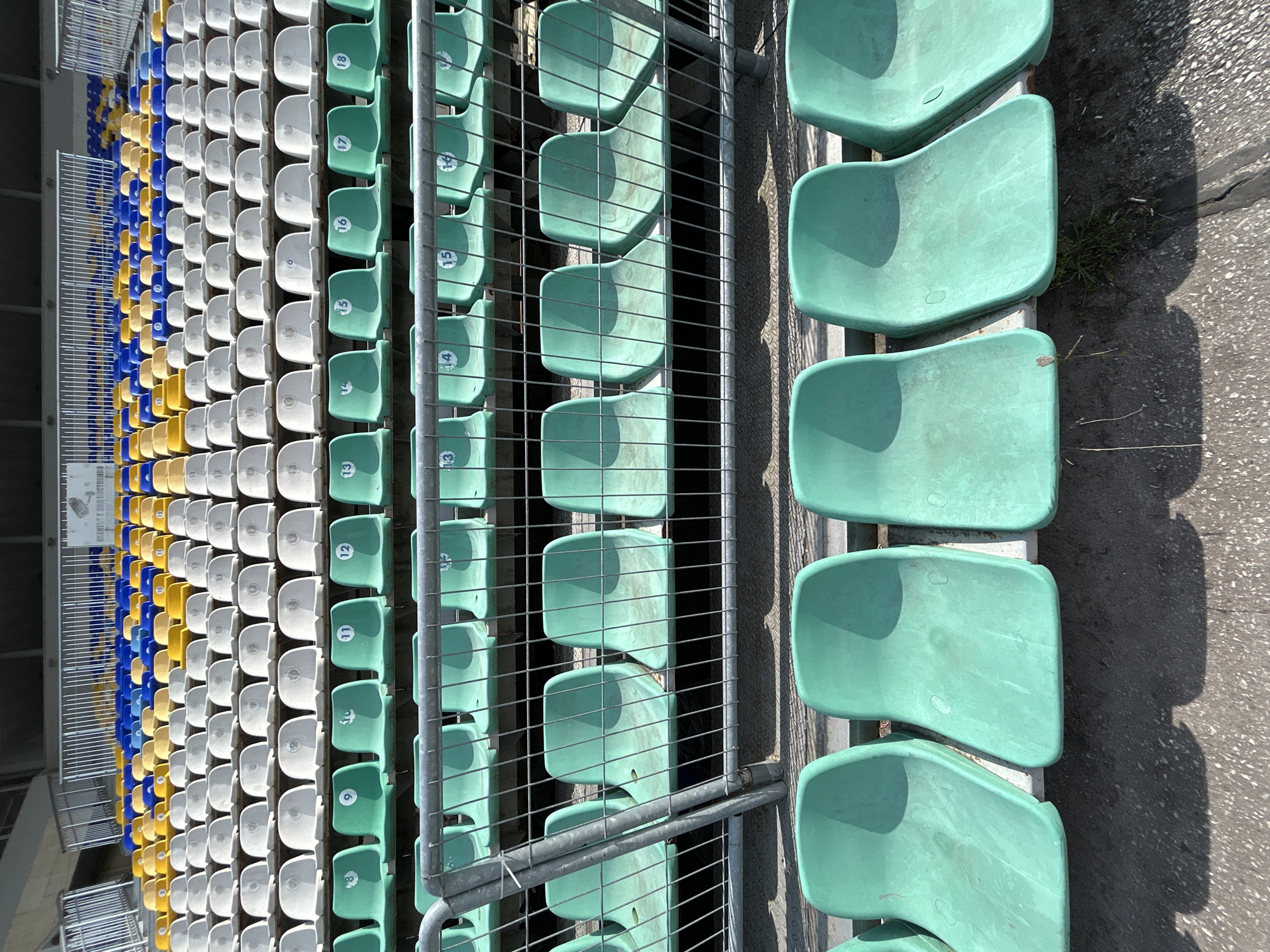 The image shows rows of stadium seats, likely taken at a sports venue in Plovdiv, Bulgaria. The seats progress in color from blue and yellow to white and then green. Many of the green seats are numbered. The seats are mounted in rows and separated by metal railings. The pavement is visible to the right of the green seats. The image appears to have been rotated 90 degrees counter-clockwise.
