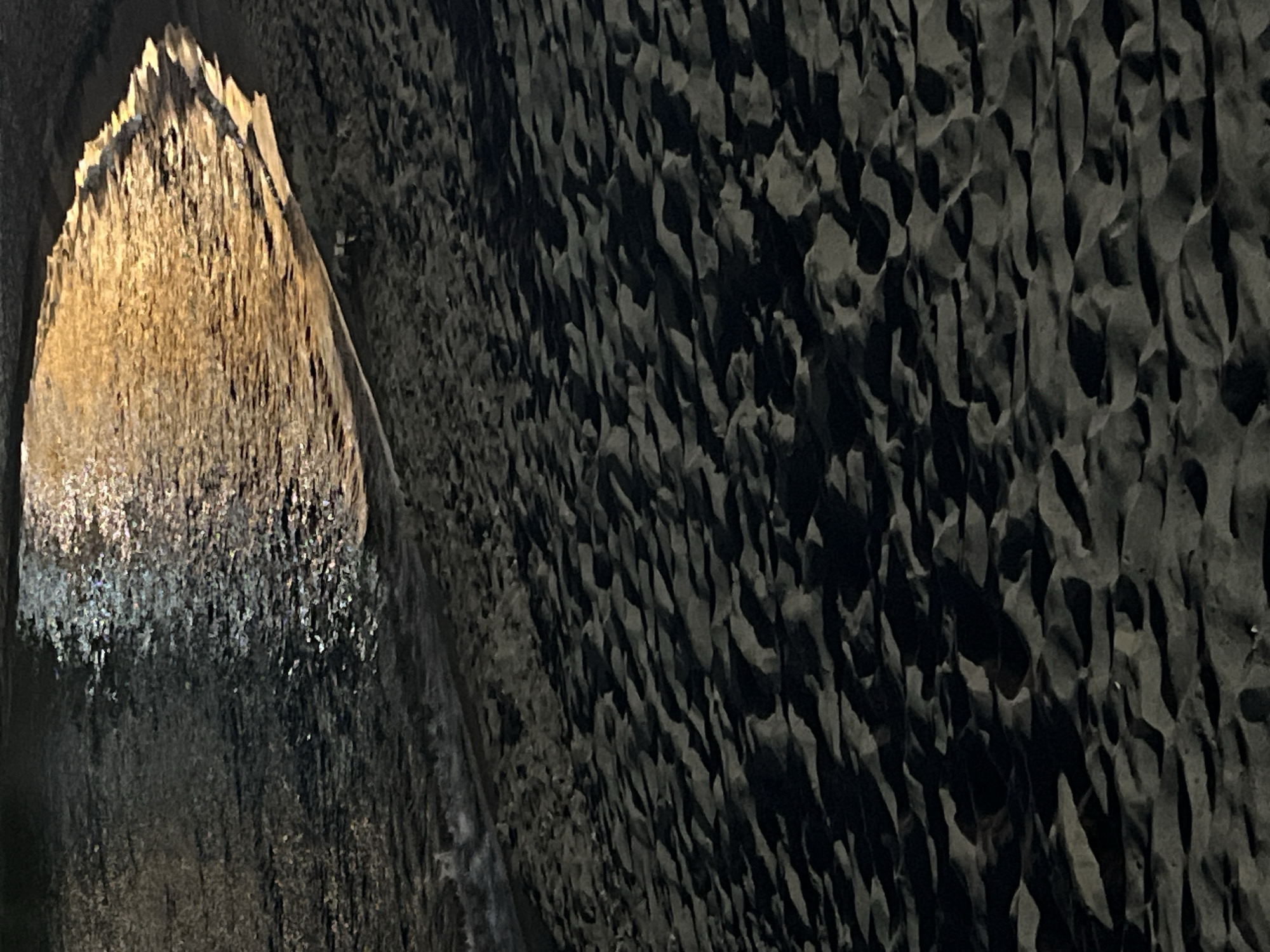 The image shows a dark sandy beach with the gentle lapping of waves. The water reflects the warm glow of artificial lights, creating a shimmering effect on the surface. The sand is rippled and textured, with shadows casting an impression of depth. The scene appears to be at night or during dusk, given the low light conditions and the prominent reflections. This setting is consistent with the beaches in Sozopol, Bulgaria, known for their accessibility and often illuminated areas after dark. There are no people or explicit objects visible in the frame, allowing the focus to remain on the natural elements of the beach and the interplay of light and shadow.