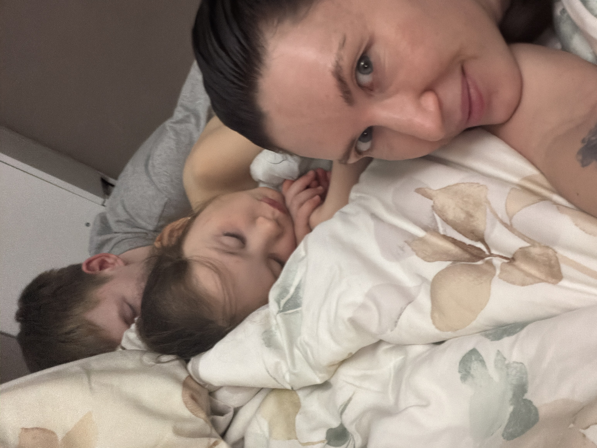 A close-up, indoor photograph depicts three individuals lying on a bed covered with a floral-patterned duvet. An adult female, with dark hair pulled back, is positioned in the foreground, looking directly at the camera with a slight smile. To her side, a young child is asleep, partially covered by the duvet. Behind the child and to the left, an adult male is also asleep. The location is presumed to be in the city of Perm, Russia. The environment appears to be a bedroom, evidenced by the bed and bedding.