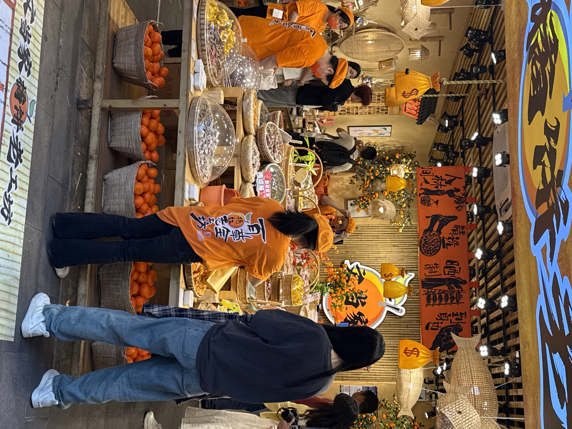 The image depicts a bustling food stall or market shop, likely indoors, characterized by traditional Chinese decor. Several individuals are present, including vendors dressed in bright orange uniforms and customers browsing the offerings.
The stall prominently features numerous woven baskets overflowing with fresh oranges, stacked on wooden shelves along the left side. In front of the oranges, clear dome-shaped containers display a variety of dried fruits or snacks. Vendors in orange shirts, some wearing face masks, attend to the display. One vendor's uniform clearly shows the text "文当家" (Wen Dang Jia) and characters that likely mean "fresh fruit" (鲜果).
Customers, including individuals wearing blue jeans and casual attire, are seen in the foreground and mid-ground, observing the products. The setting includes bamboo-like wall panels and decorative elements such as woven lanterns, some shaped like money bags marked with a dollar sign ($). Overhead, track lighting illuminates the display, and string lights add to the festive atmosphere. A large circular sign with an orange background, featuring Chinese characters and a fruit motif, is visible on the right side of the stall. A red banner hanging vertically displays text including "永川黑鸭" (Yongchuan Black Duck), "精" (refined), "精装坊" (boutique/fine decoration workshop), and "当家香饼老工坊" (Master's fragrant cake old workshop), suggesting a variety of traditional goods are available or promoted in the area. The overall impression is a lively market environment, rich with local flavors and cultural elements, likely in Yuzhong District, China.