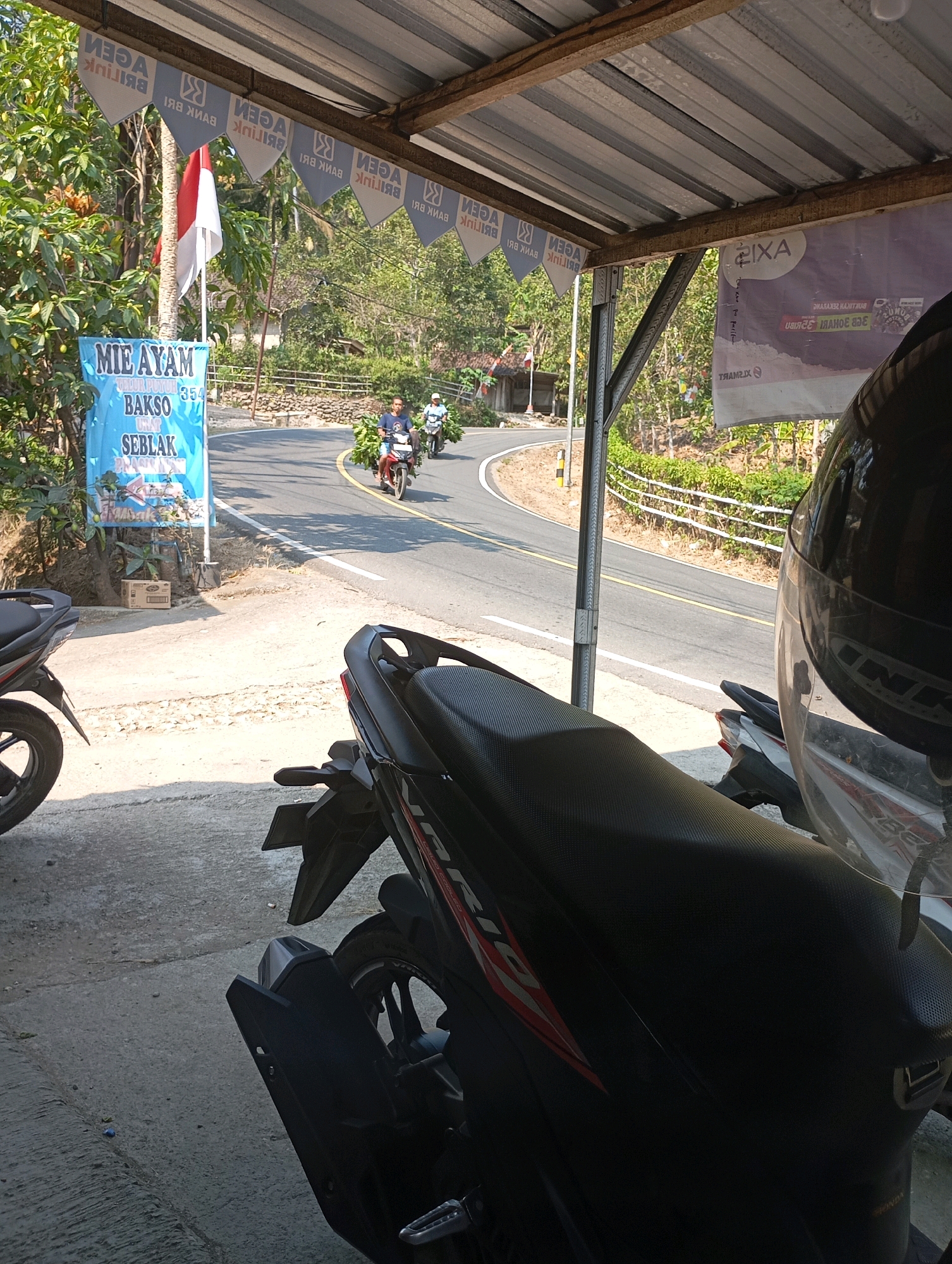 The image depicts a roadside scene in Ketepung, Indonesia, likely a small town or village. In the foreground, two motorcycles are parked, one in partial view and the other dominating the lower right of the frame. A black motorcycle helmet rests on the seat of the visible motorcycle. A blue banner, featuring text in Indonesian including "MIE AYAM", "BAKSO", and "SEBLAK", is prominently displayed on the left, suggesting a food stall or restaurant. The background shows a winding road with two men on motorcycles traveling along it. The man on the left is carrying what appears to be a large bunch of green foliage. The road is flanked by trees and vegetation, with a white fence line visible further up the hill. The scene is covered by a metal roof supported by a metal pole, indicating a covered area, possibly a shelter or extension of a building. Indonesian flags are visible hanging from the roofline, interspersed with triangular pennants bearing the text "AGEN BANK BRI". The overall impression is one of a typical Indonesian roadside establishment on a sunny day.