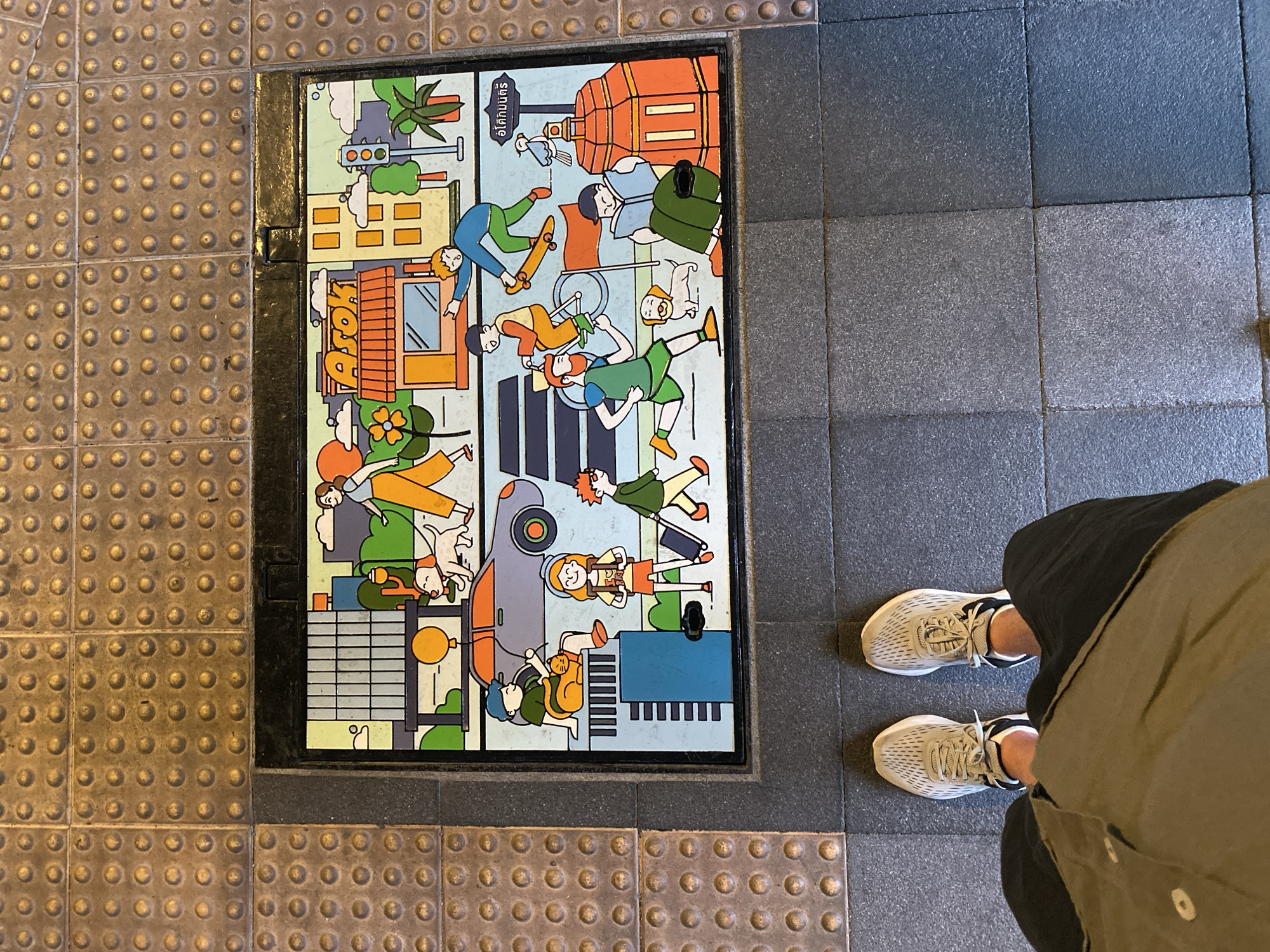The image shows a ground-level view of a textured public walkway in Bangkok, Thailand. A rectangular metal plate is set into the pavement, adorned with a colorful cartoon-style illustration depicting various people and activities within a city. The artwork includes buildings, trees, vehicles, and pedestrians engaged in everyday life, like skateboarding, cycling, and walking pets. The inscription "อิโคมิวเซียม" is visible on the top right of the plate. Adjacent to the illustrated plate are two types of tactile paving, one with raised dots and the other with a pattern of grey squares. A person is standing to the right of the metal plate, wearing light-colored running shoes, dark trousers, and a button-down shirt or jacket.