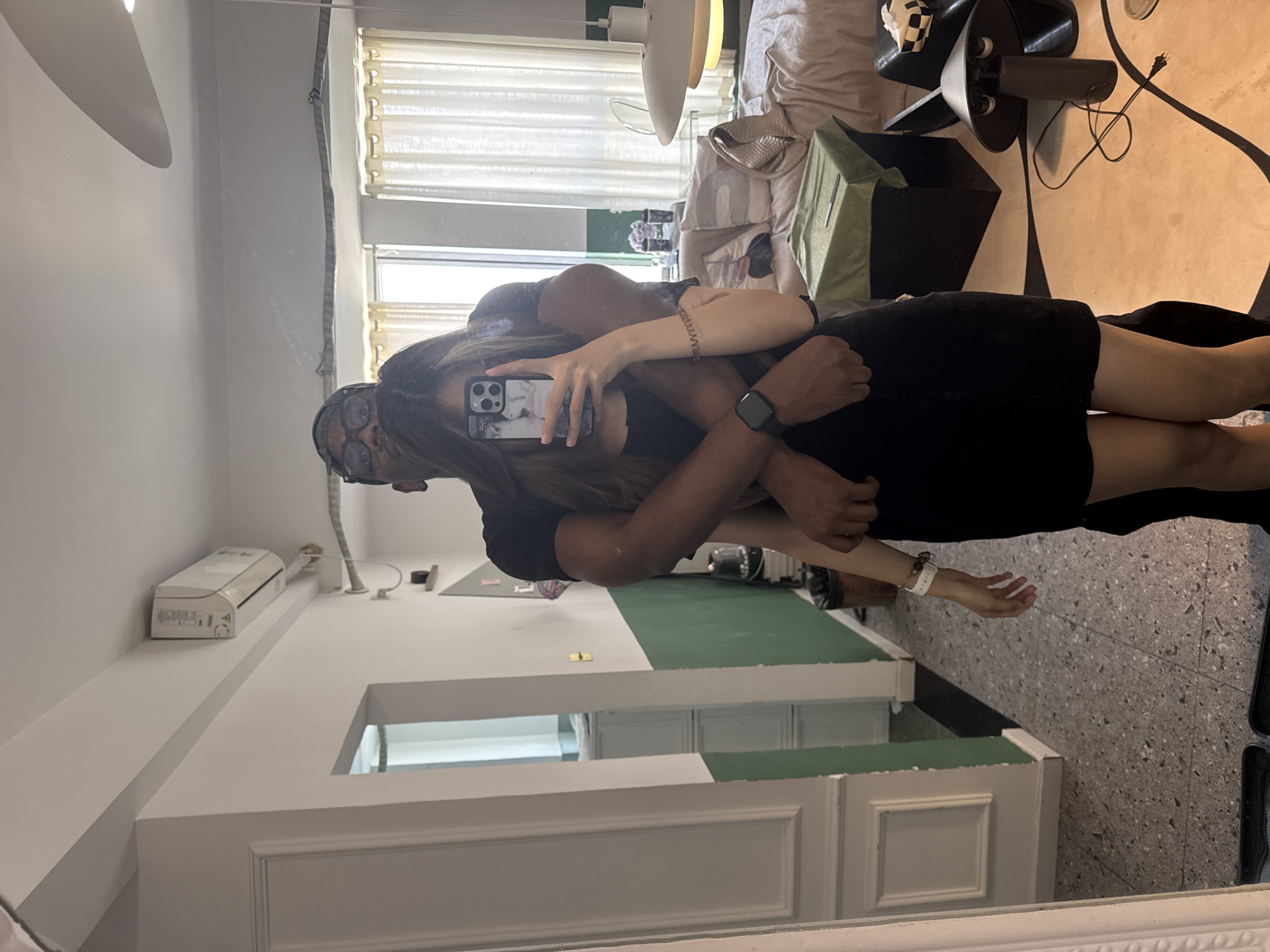 This mirror selfie captures a couple embracing in what appears to be a modern indoor setting in Liangjiang, China. The image is a reflection in a large mirror featuring a white, decoratively molded frame.
The subjects are a man and a woman. The woman, with long dark hair and glasses, is holding a smartphone with a marble-patterned case, taking the picture and partially obscuring her face. She is wearing a dark dress or top, and a light-colored watch or bracelet is on her left wrist. The man, with a darker complexion and glasses, is wearing a dark short-sleeved shirt and a smartwatch on his left wrist. He is embracing the woman, his arm around her, and his head resting near hers, suggesting an affectionate and intimate moment.
The reflected room features white walls, with a section of the lower wall painted dark green. The floor is light grey with a speckled, possibly terrazzo, texture. Natural light illuminates the room through windows fitted with light-colored blinds or curtains. Visible furnishings and objects include a white air conditioning unit mounted high on a wall, a long white shelf or counter, a modern ceiling light fixture, and a sofa or bed with light-colored cushions. A black side table holds a dark, multi-legged object (possibly a fan or lamp) and a small checkerboard-patterned item. A green fabric storage bin is also visible, along with some electrical cables on the floor. The overall scene suggests a comfortable domestic or hotel environment, captured during the daytime. No discernible text is visible in the image.