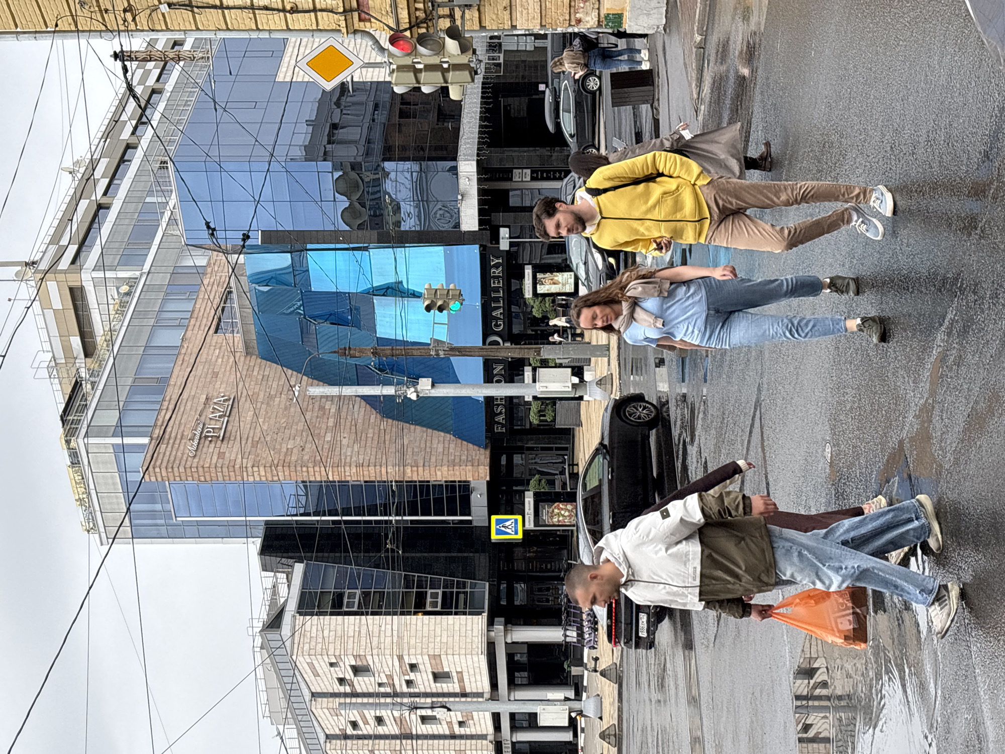 This is a wide shot, eye-level, outdoor photograph of a street scene in Nizhny Novgorod, Russia. The weather appears to be overcast and the pavement is wet, suggesting it has recently rained or is still raining. Several people are walking across a street intersection.
In the foreground, a man wearing a white and olive green jacket and blue jeans walks from left to right, carrying an orange plastic bag in his left hand. He is looking to his right. Beside him and slightly behind, a woman in a light blue t-shirt and jeans walks from left to right, with a beige scarf around her neck. She is looking forward. Behind them and to the right, a man in a bright yellow hooded jacket and brown pants walks from left to right, looking forward. He is holding a dark object in his right hand. Other pedestrians and a dark colored car are visible in the background.
The background features a modern building complex with large glass windows and brick accents. A sign on the building reads "FASHION GALLERY". Traffic lights are visible, with one showing a green light and another showing a red light. Overhead wires crisscross the sky.