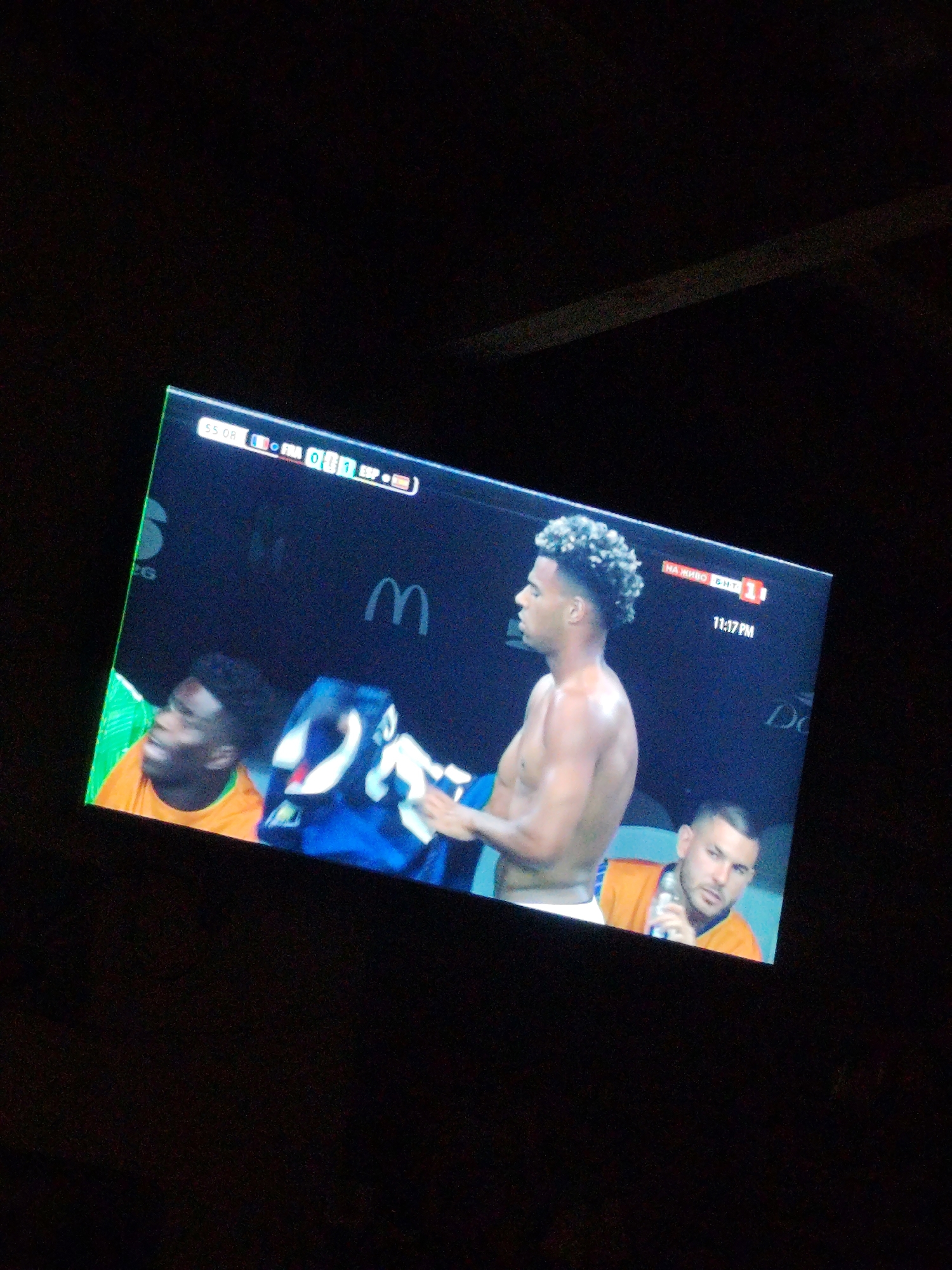 The image shows a television screen displaying a soccer match. The score is 0-1. The screen shows three male soccer players and a logo for "McDonald's". The screen also displays "11:17 PM" and "НА ЖИВО БНТ1". The TV is viewed at an angle in a dark room. The image appears to be taken in Prolesha, Bulgaria.