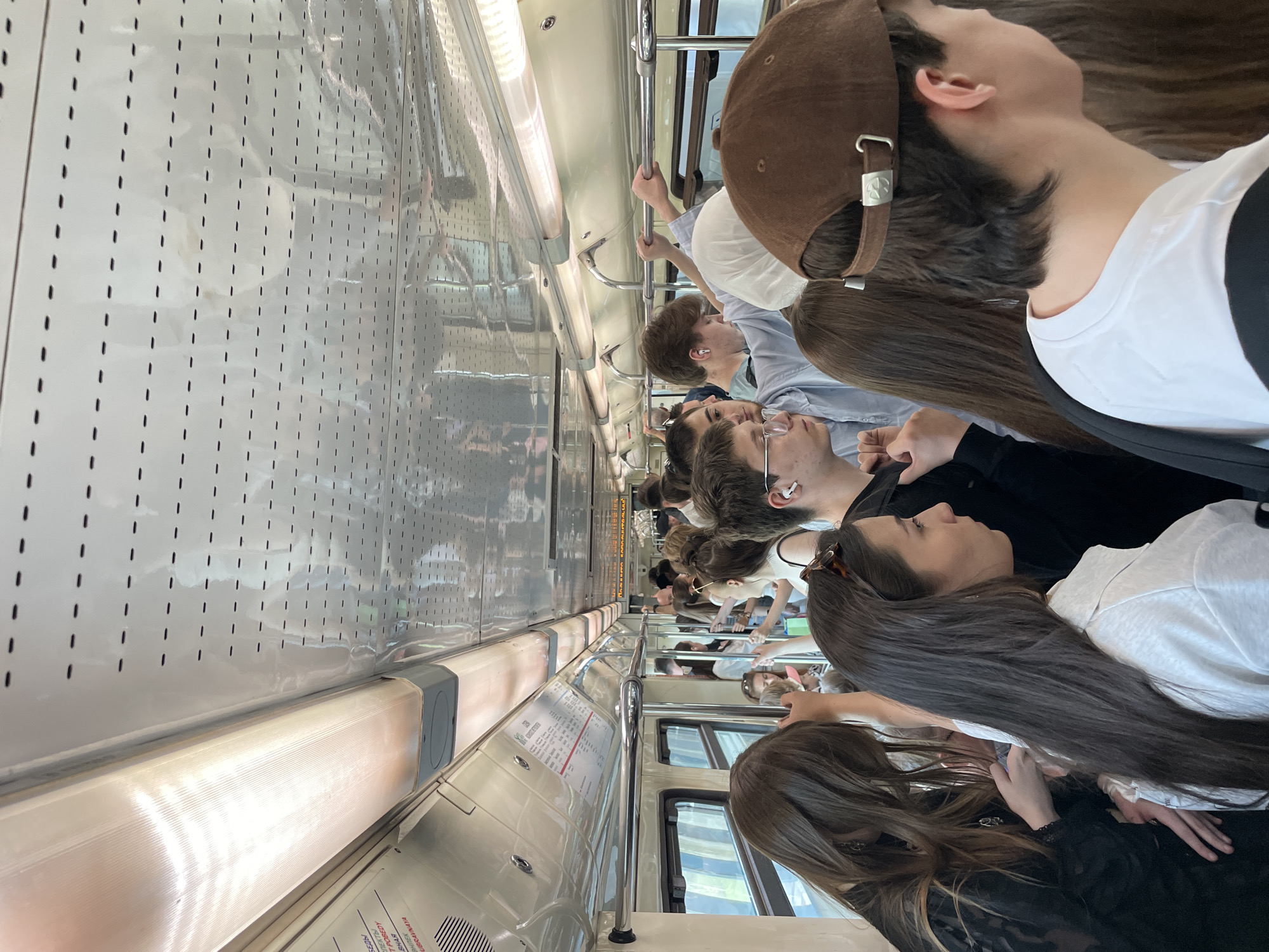 The image depicts the interior of a crowded train car, likely a subway or commuter train, filled with passengers. The interior of the car is sterile with white perforated panels and stainless steel handrails. The passengers are a diverse group of people standing close together, occupying the limited space. Some are looking forward, while others appear to be engaged with their phones or other personal devices. The overall impression is one of a typical scene during peak travel hours on a public transportation system.