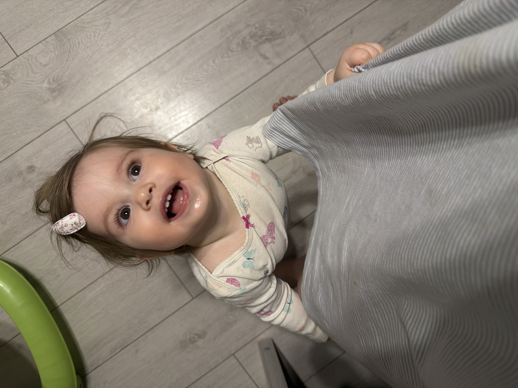 A young child in a patterned onesie stands on a light-colored wooden floor, holding onto a blue and white striped curtain. The child has light brown hair, a floral clip in their hair, and is looking up and smiling with an open mouth. A green, inflatable banana-shaped toy is visible in the lower left corner of the frame.