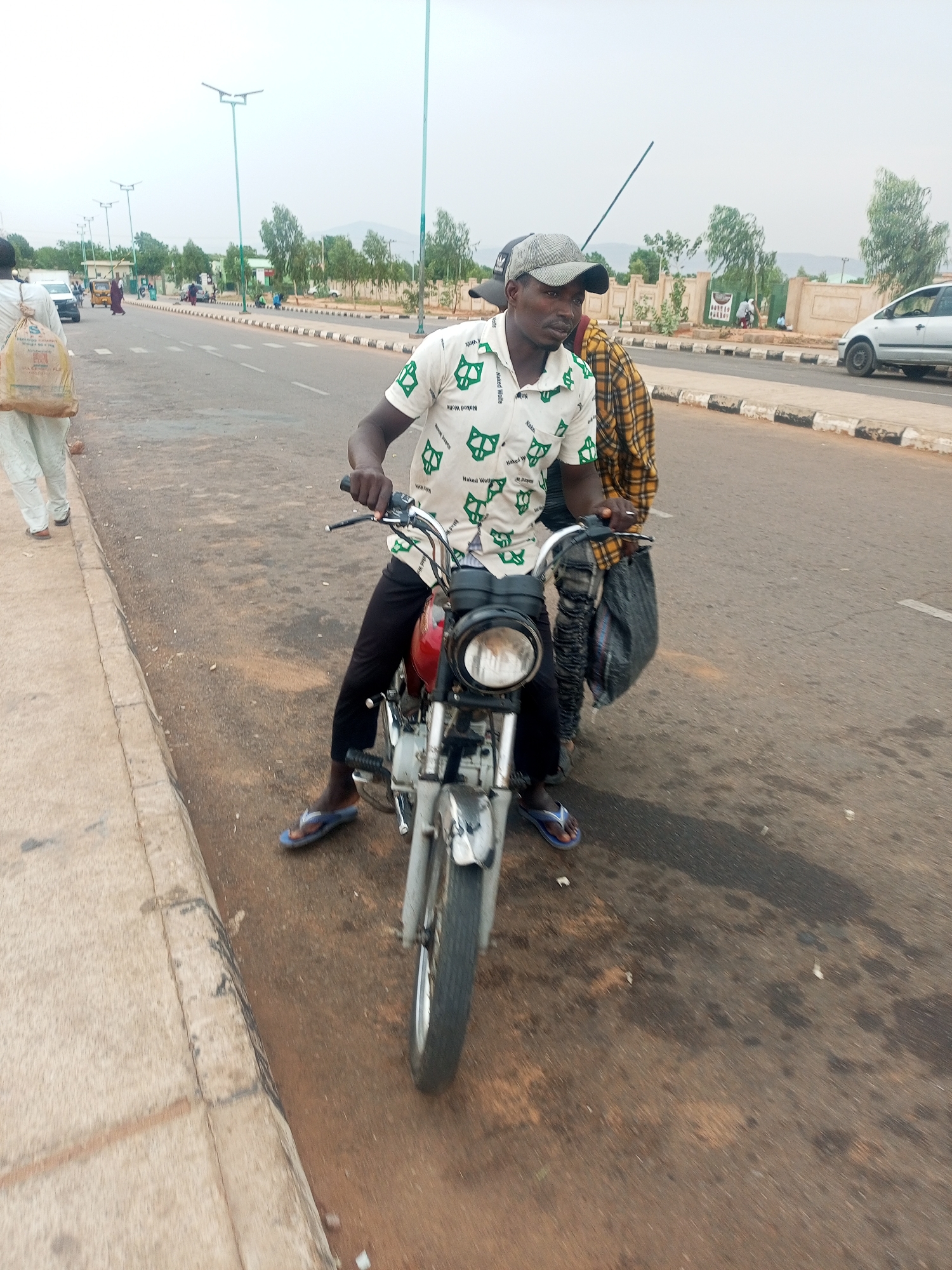 A man wearing a white shirt with green, patterned designs, black pants, and blue sandals is seated on a red and silver motorcycle at the edge of a paved road. He also wears a gray and black cap. A second man, partially visible behind him, wears a yellow plaid shirt and carries a black bag. The location is Gombe, Nigeria. In the background are trees, lampposts, a white vehicle, and other people. The road has a raised curb with black and white painted sections.