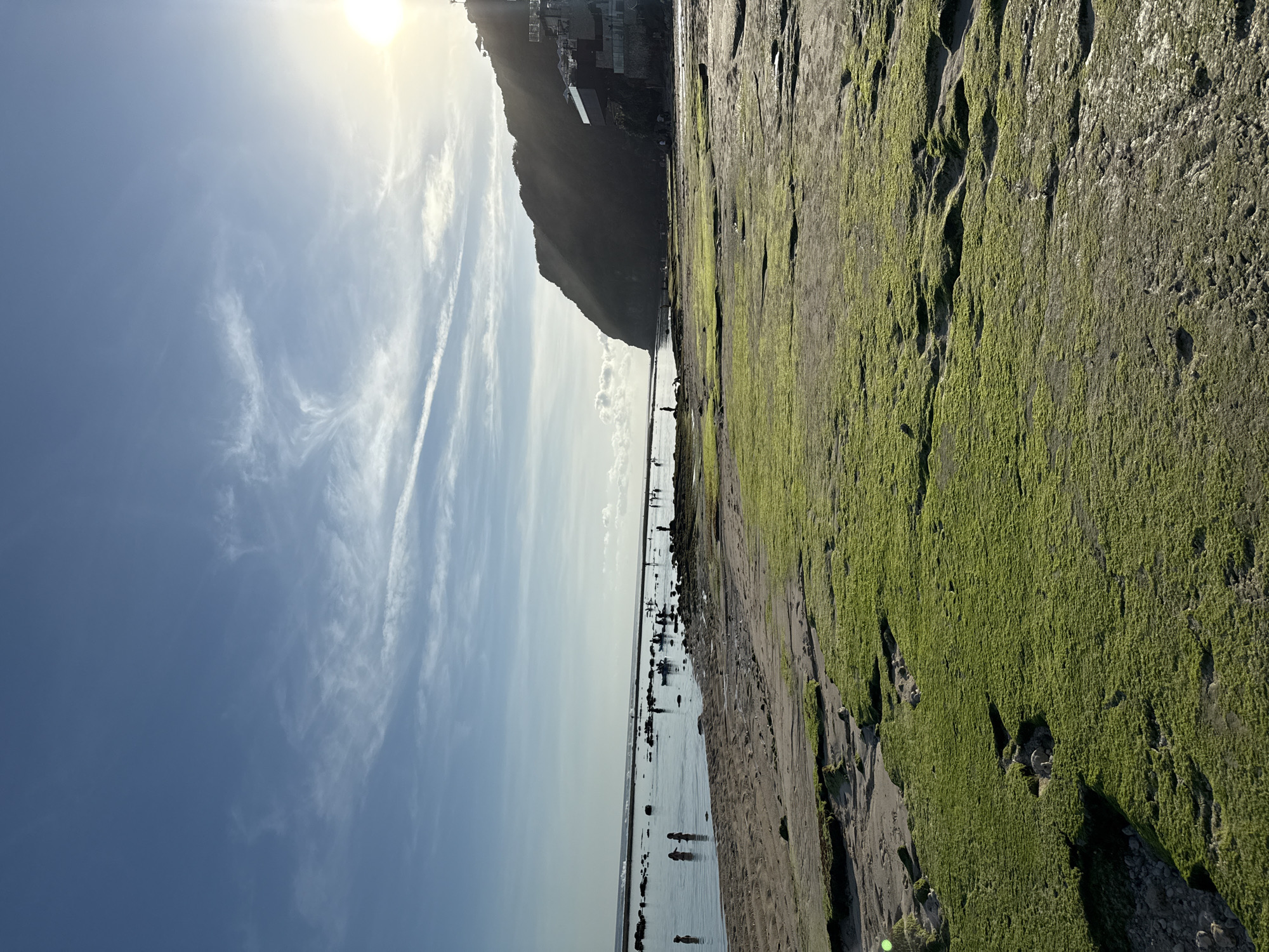 The image shows a coastal scene in Kuta Selatan, Indonesia. The photograph is oriented vertically. The sky is a light blue with wispy white clouds in the upper left. The sun is at the top center, creating glare. A dark, rocky headland dominates the center of the image. Its base merges with a sandy beach populated by multiple individuals. Some appear to be wading in the shallow water. The right side of the image features a green, moss-covered rocky surface. There is no obvious human activity on the moss.