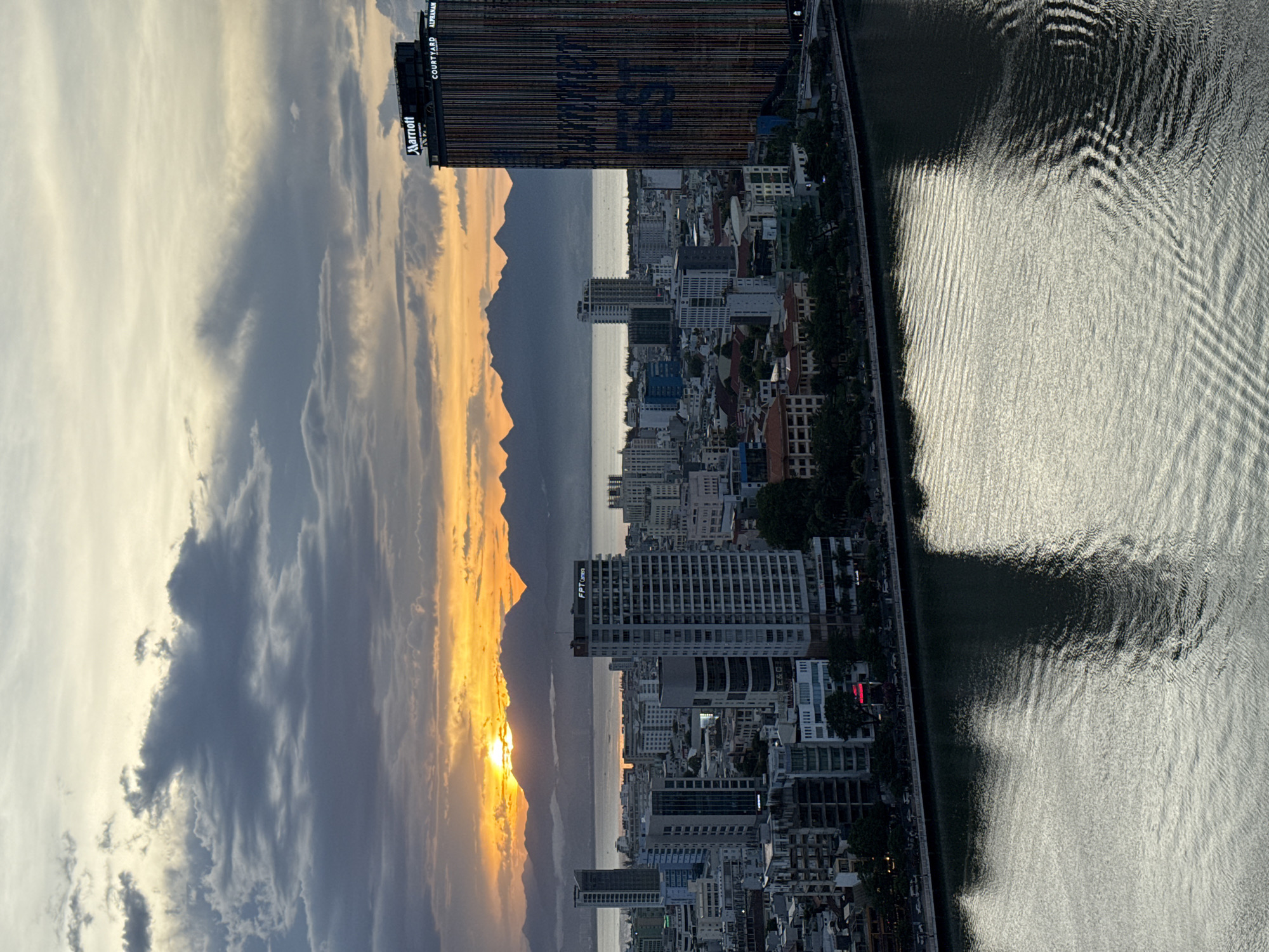 An aerial view captures a city skyline at sunset, with the Han River in the foreground. The water reflects the overcast sky and the city lights. Buildings of varying heights dominate the cityscape, including the Marriott and Courtyard by Marriott hotel with its distinctive vertical lines and large text on its facade. A range of mountains forms the distant horizon. The setting sun casts a warm glow on the clouds and the mountain range, creating a dramatic sky. The scene depicts Đà Nẵng, Vietnam.