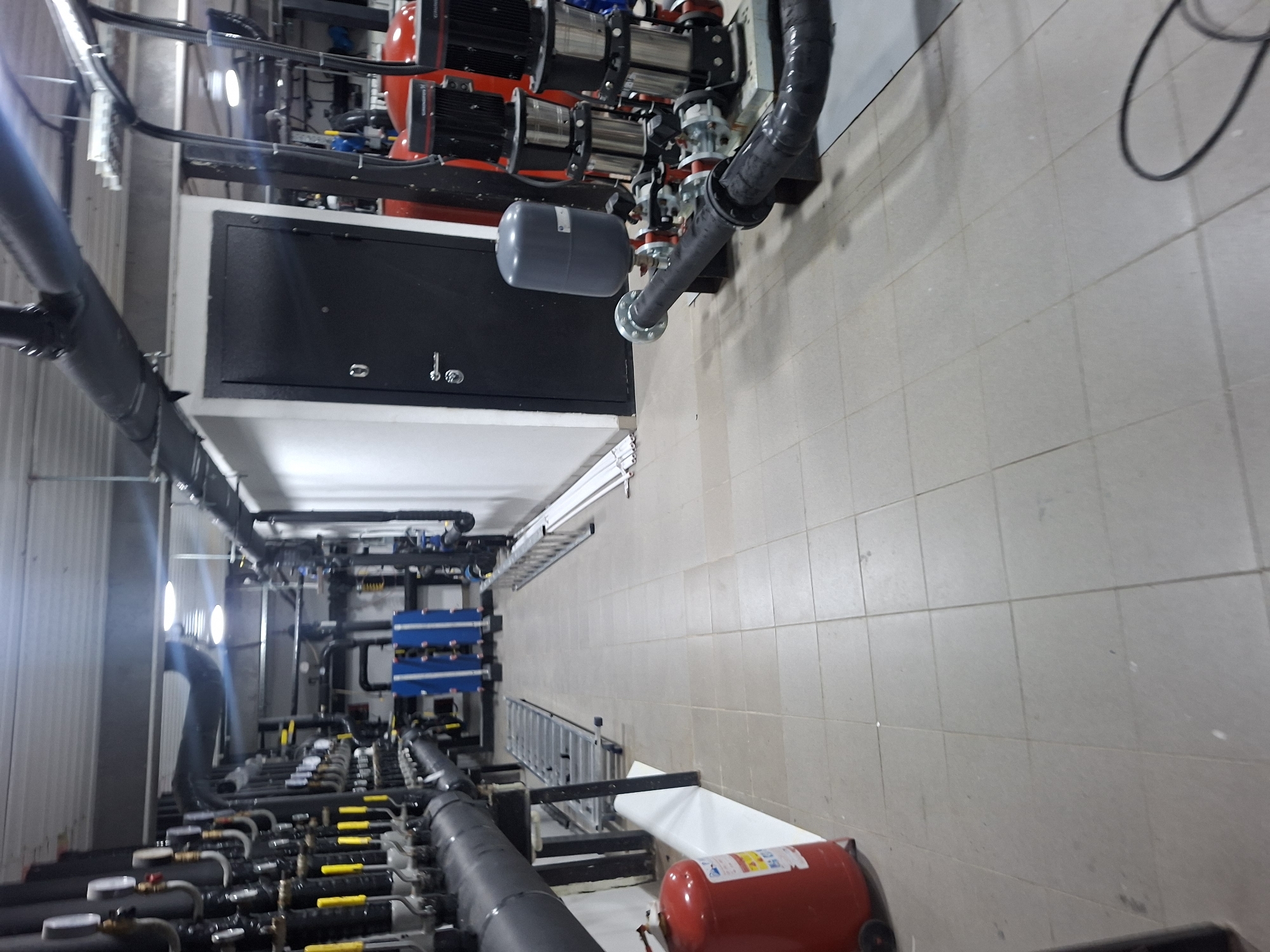 The image depicts a mechanical room, likely within an industrial or commercial facility, located in Kokshetau, Kazakhstan. The room contains complex piping systems, pumps (Grundfos), valves (with yellow handles), and electrical equipment. A grey expansion tank is visible. There is a dark grey metal control box with handles. A red fire suppression tank rests on the floor. A ladder and what appears to be a white tub are also present. The floor is covered in light colored tiles. There is an absence of human presence and a focus on the installed machinery and utilities.