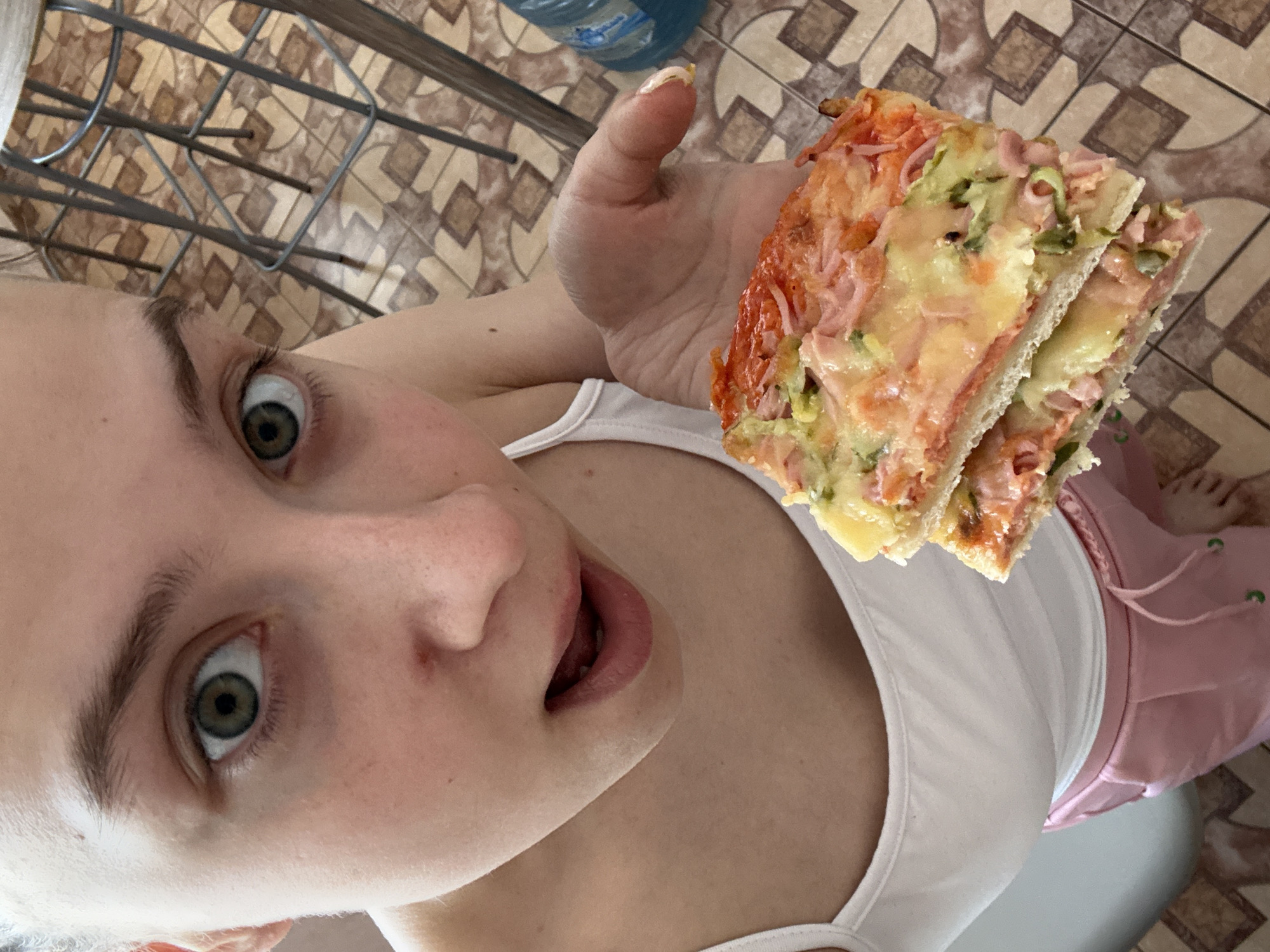 A low-angle shot captures a woman with light skin and light-colored hair, wearing a white tank top and pink shorts. Her eyes are wide and directed towards the camera, and her mouth is slightly open. She holds a stack of two square-cut pieces of pizza in her right hand. The pizza appears to have toppings of cheese, ham, and green vegetables. The background includes a tiled floor with a geometric pattern, and a blue water bottle is partially visible at the top of the frame. The location is in the city of Прикамский, Russia.