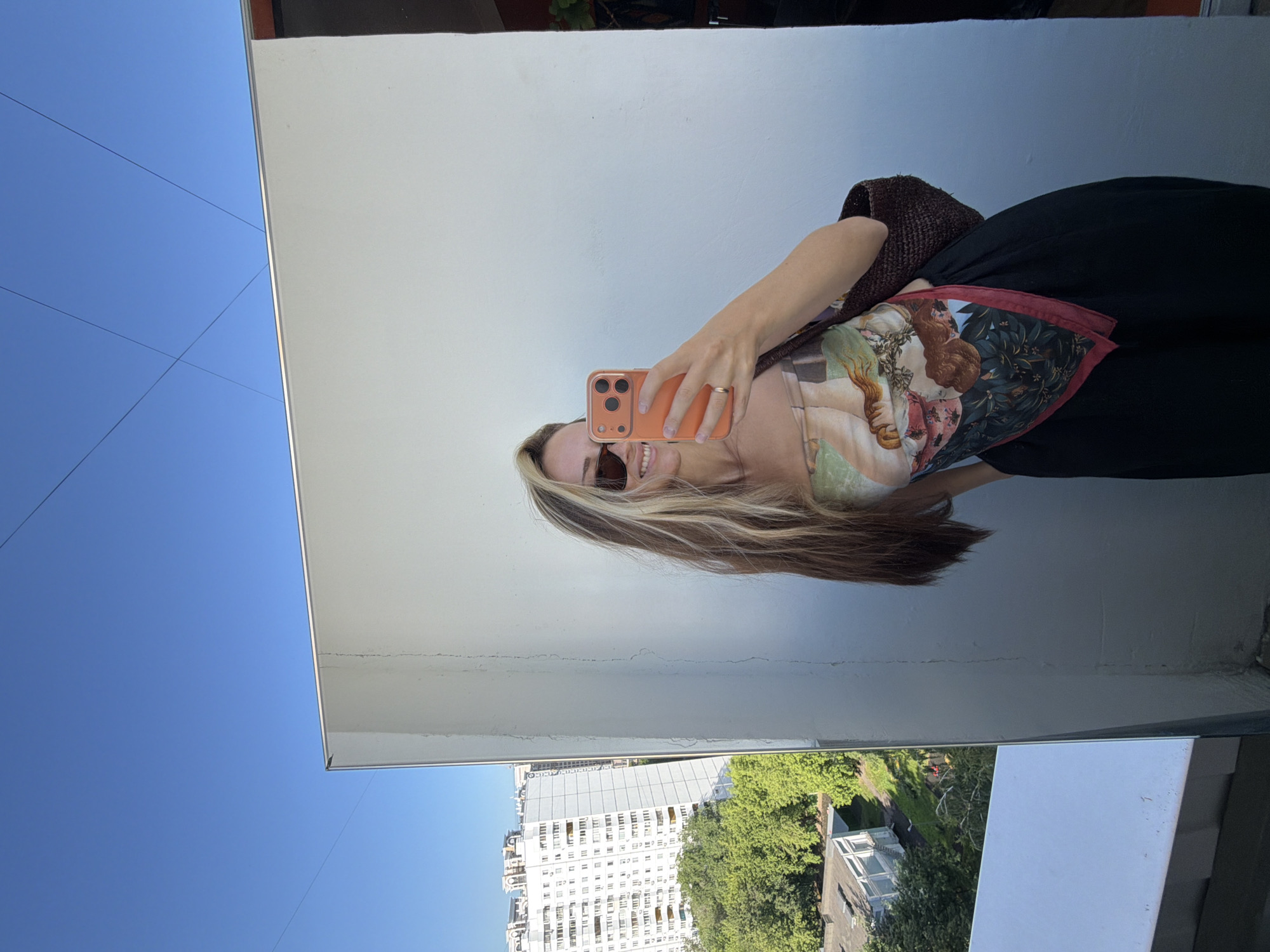In Khimki, Russia, a young woman with long, blonde hair and sunglasses poses for a selfie against a plain wall, reflecting an urban cityscape. She wears a patterned bandana top and black pants, with a woven bag slung over her shoulder. The reflection in the mirror reveals a tall, white apartment building and lush green trees, suggesting a residential area with a view. The sky is a clear, bright blue.